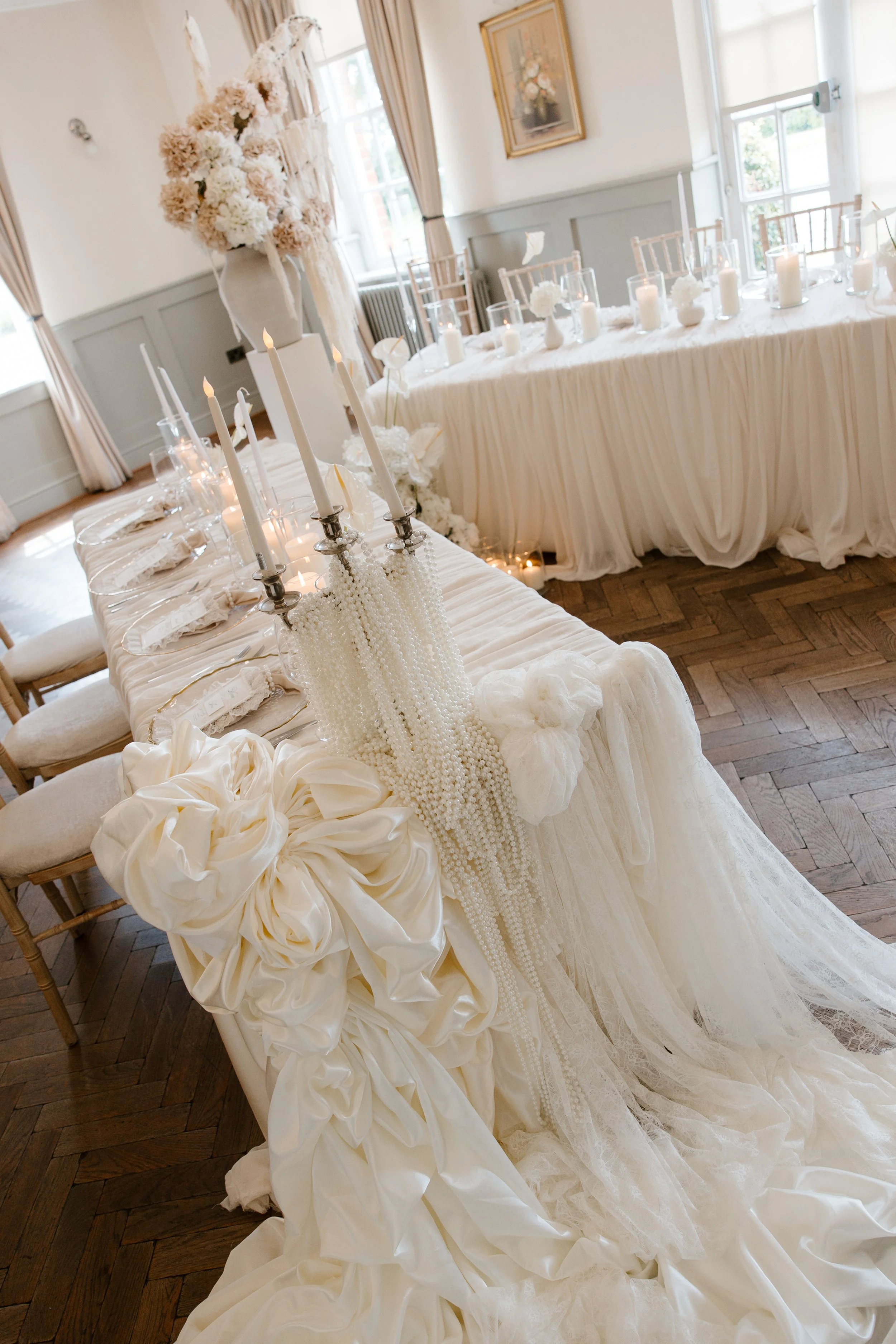 Elegant wedding reception table setup with white fabric tablecloths, tall white candles, floral centerpieces, and decorative elements.