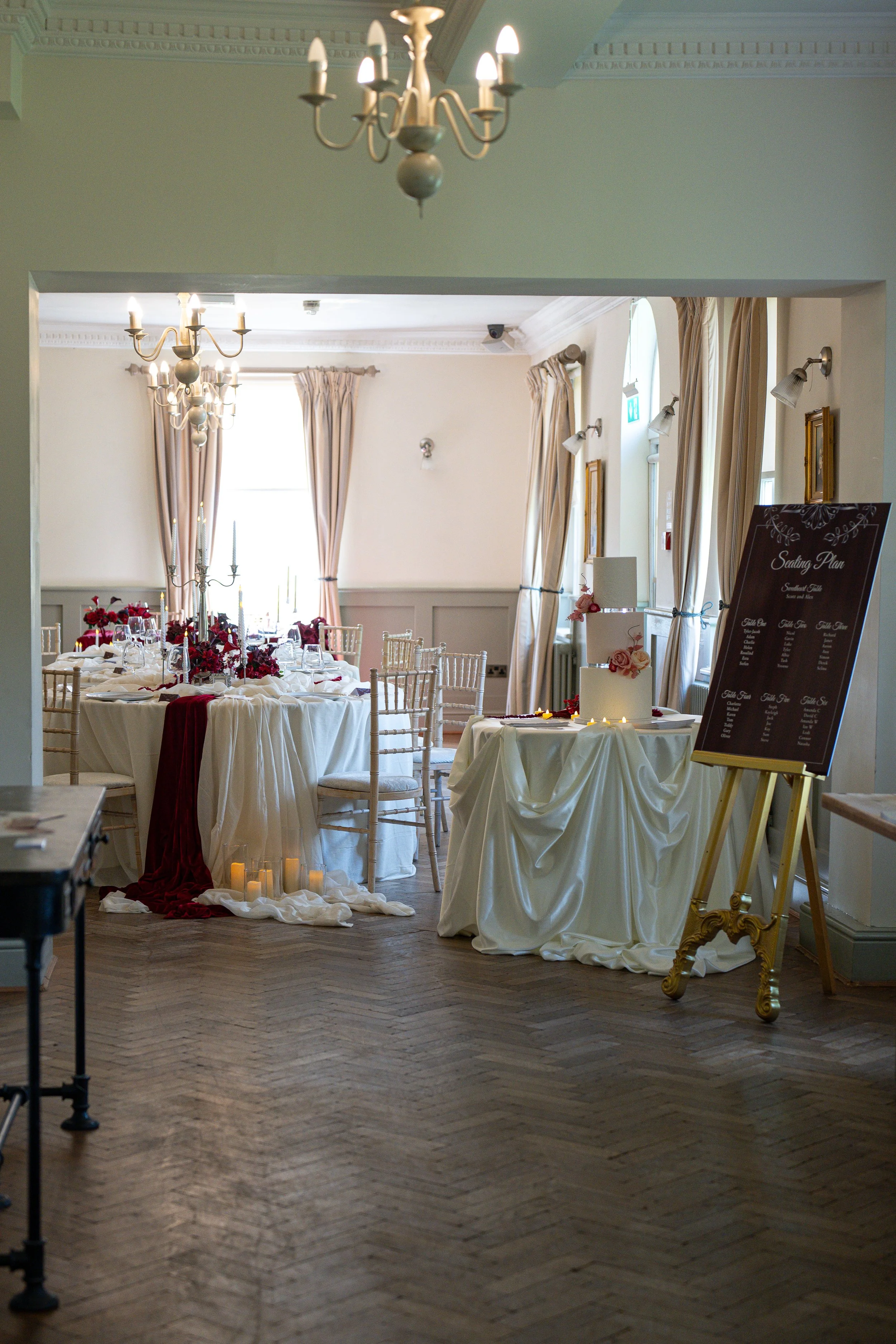 Elegant wedding reception setup with decorated tables featuring red and white floral centerpieces, candles, and white tablecloths, set in a well-lit room with chandeliers, tall windows with beige curtains, and a wedding cake on a side table.