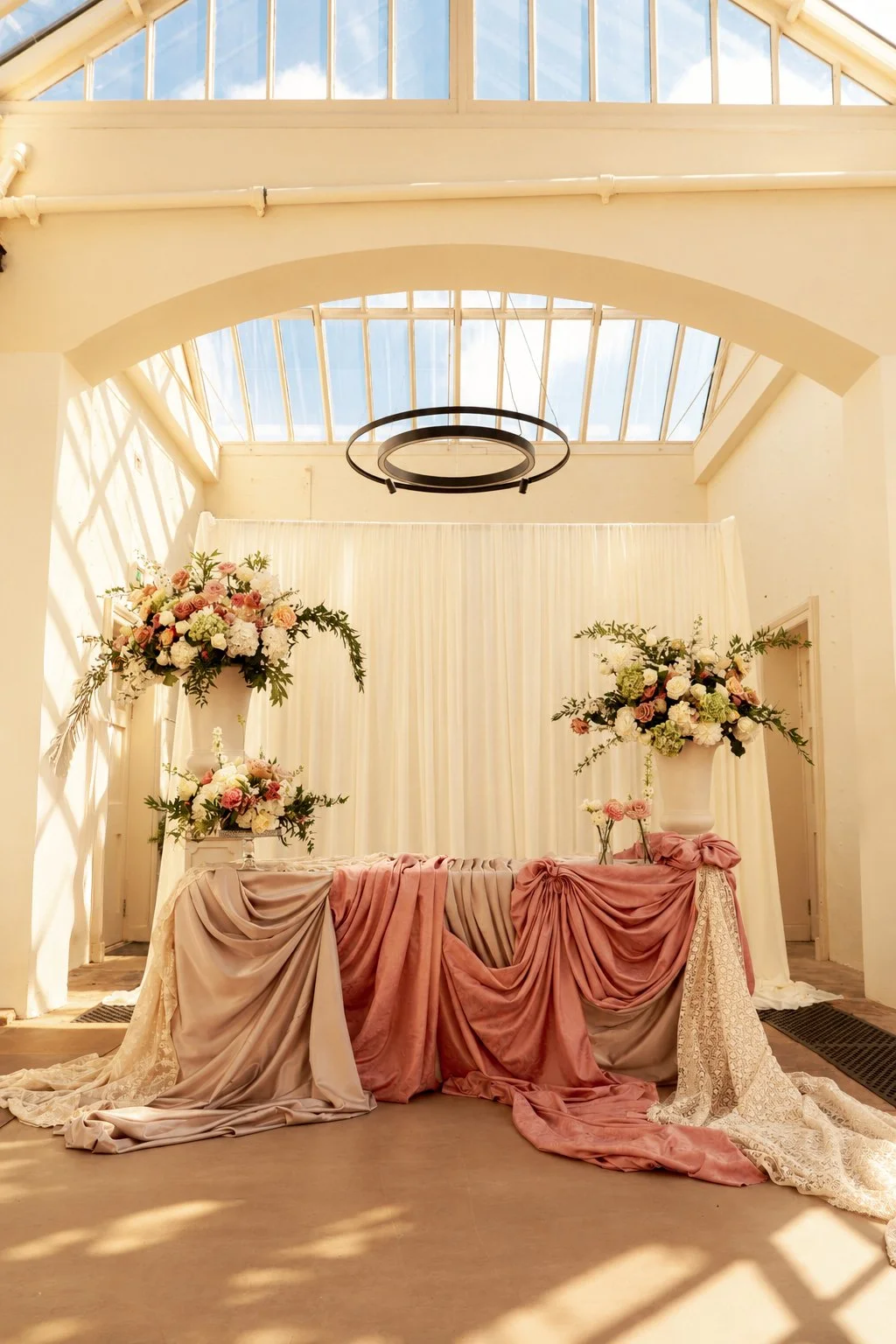 Elegant room decorated for wedding with pink and cream draped fabric and large floral arrangements of white, pink, and green flowers, set in tall white vases, in front of a cream-colored curtain and natural sunlight from a vaulted glass ceiling.