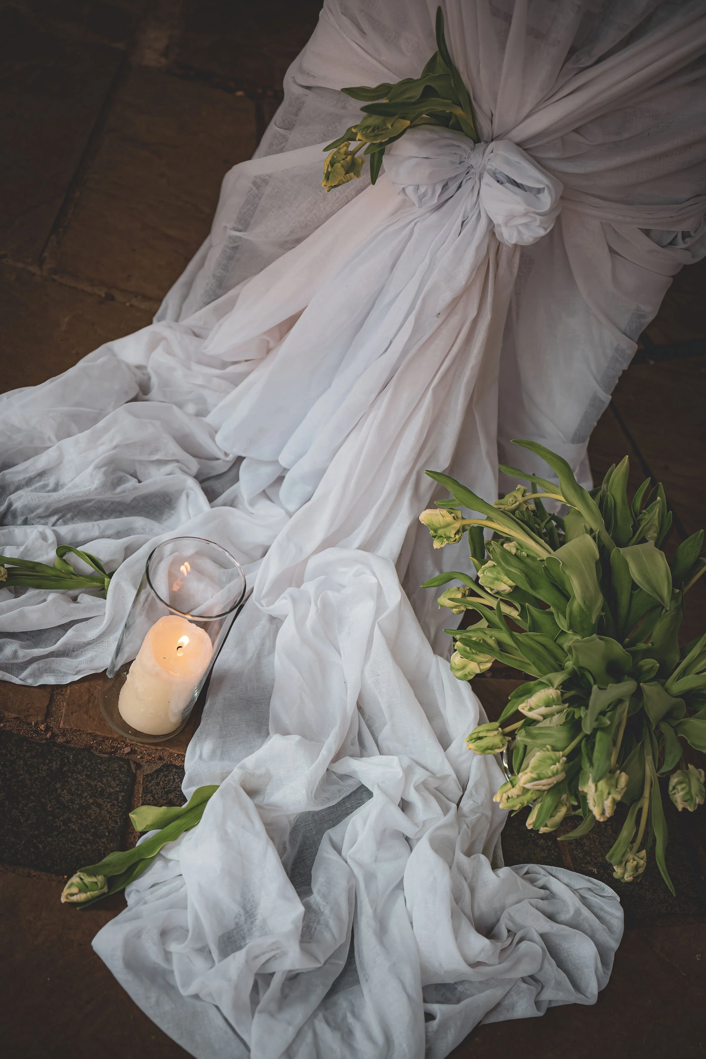 A white fabric is draped and tied in a knot on a dark floor, with green foliage and white flowers nearby, and a lit candle in a glass holder.