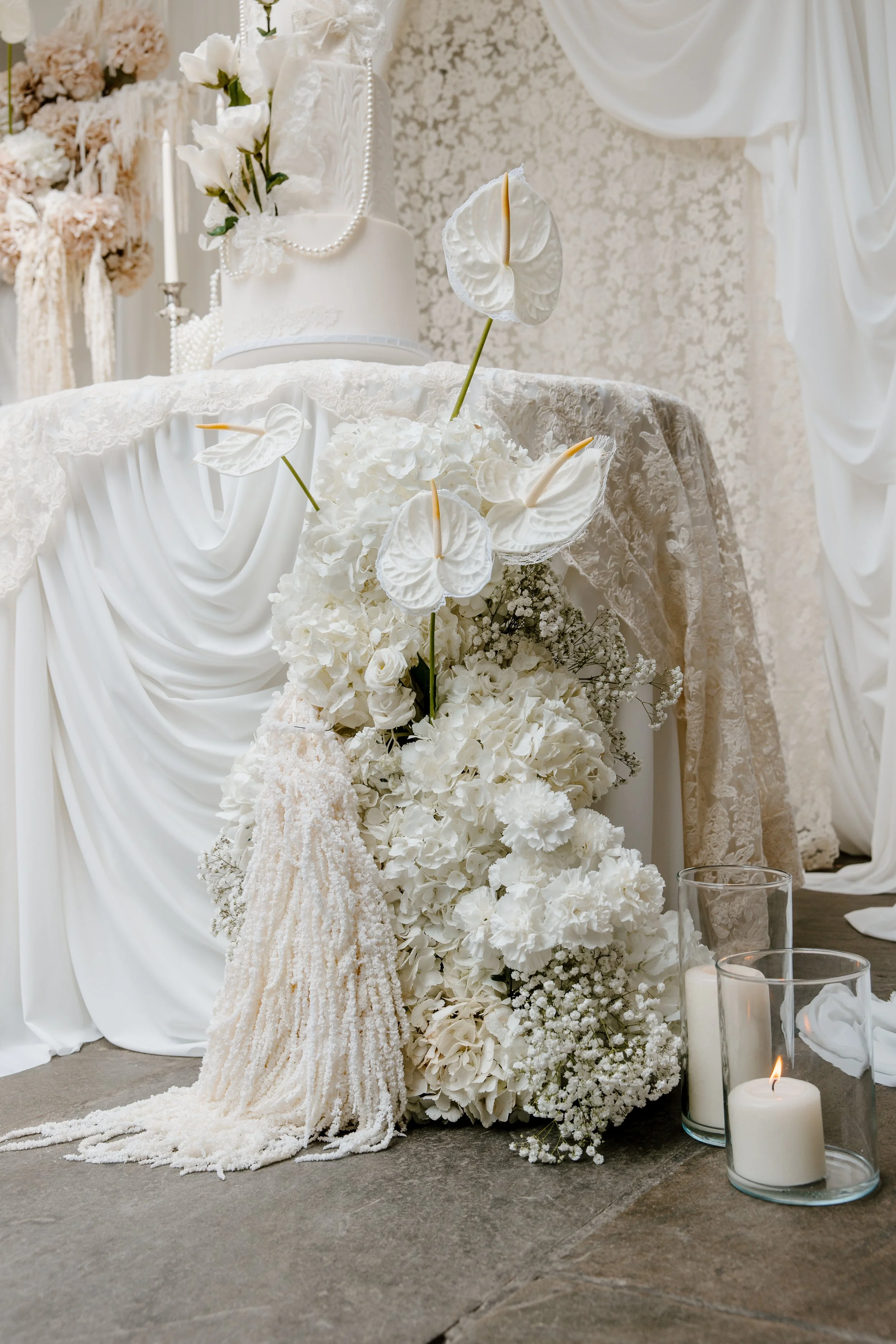 A white wedding cake with floral decorations, surrounded by white flowers and candles, on a table draped with white fabric in a wedding setting.
