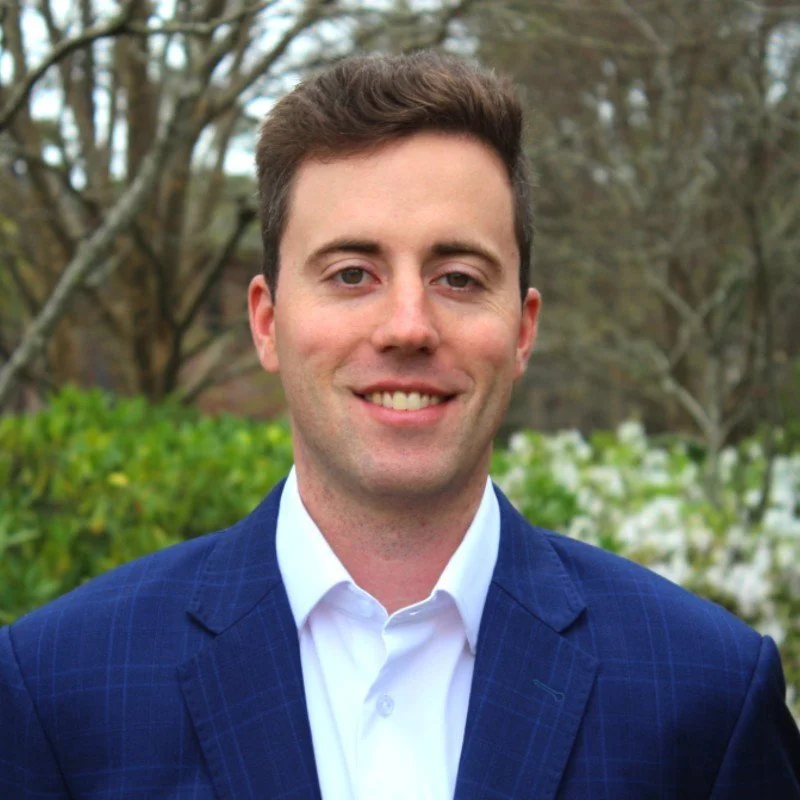 Portrait of a smiling man in a blue suit outdoors with trees and greenery in the background.