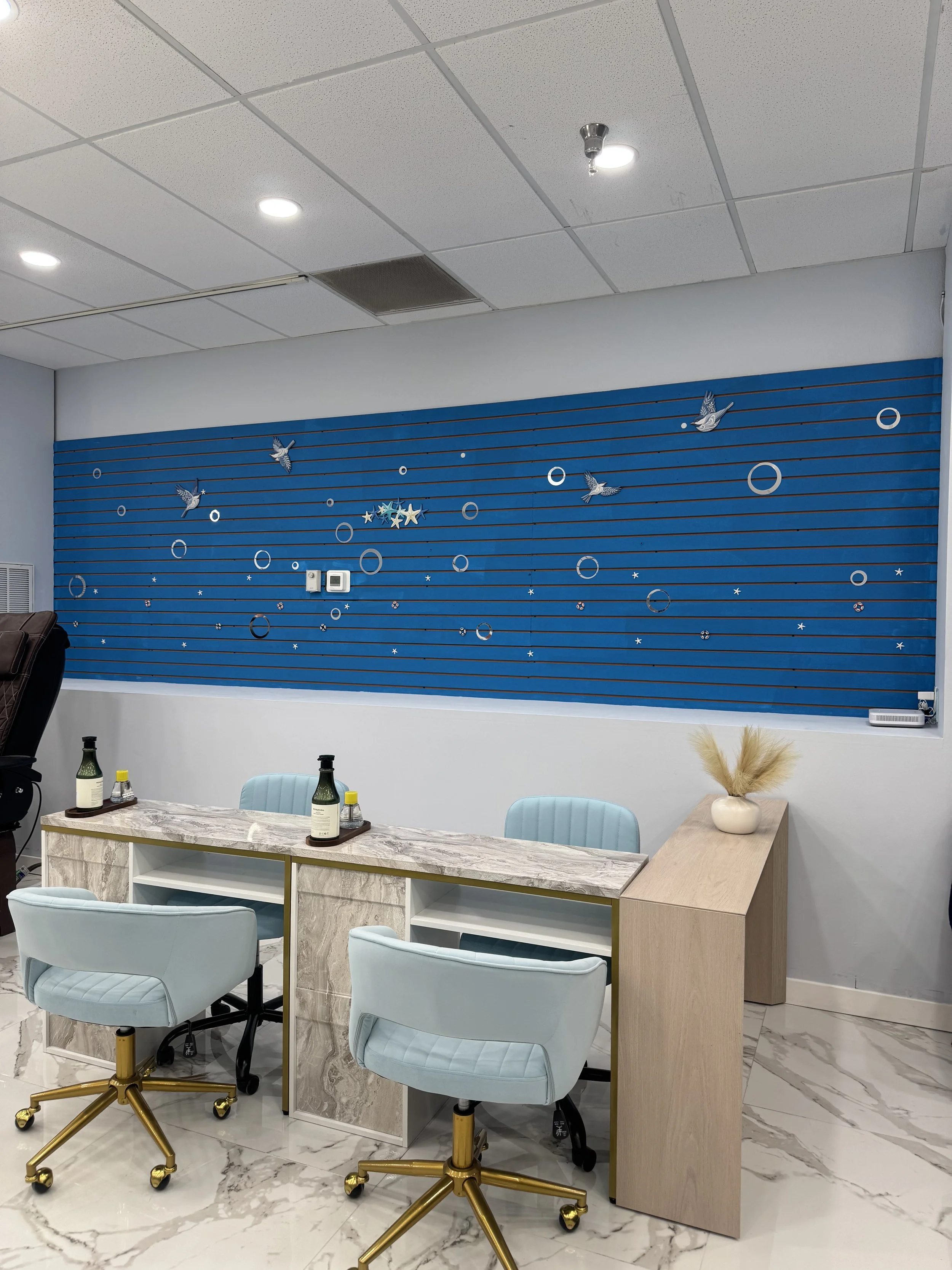 Office meeting room with a marble-style table, light blue chairs with gold bases, a blue wall with decorative hummingbird and circle designs, and a beige side table with a vase of dried flowers.