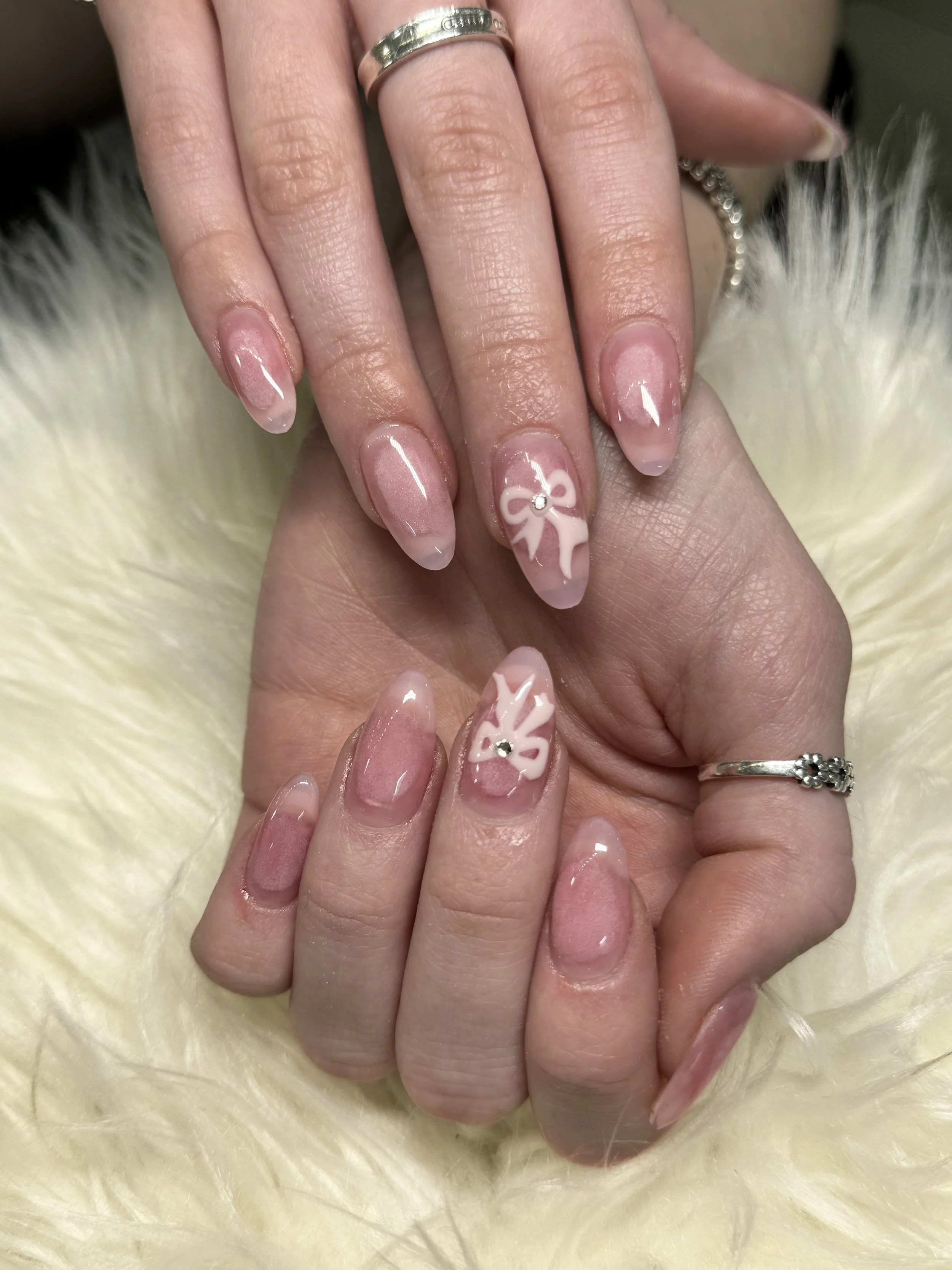 Close-up of a person's hand with manicured nails, featuring pink and white nail art with a bow design and rhinestone accents. The person is wearing rings, and the background shows a soft, furry surface.