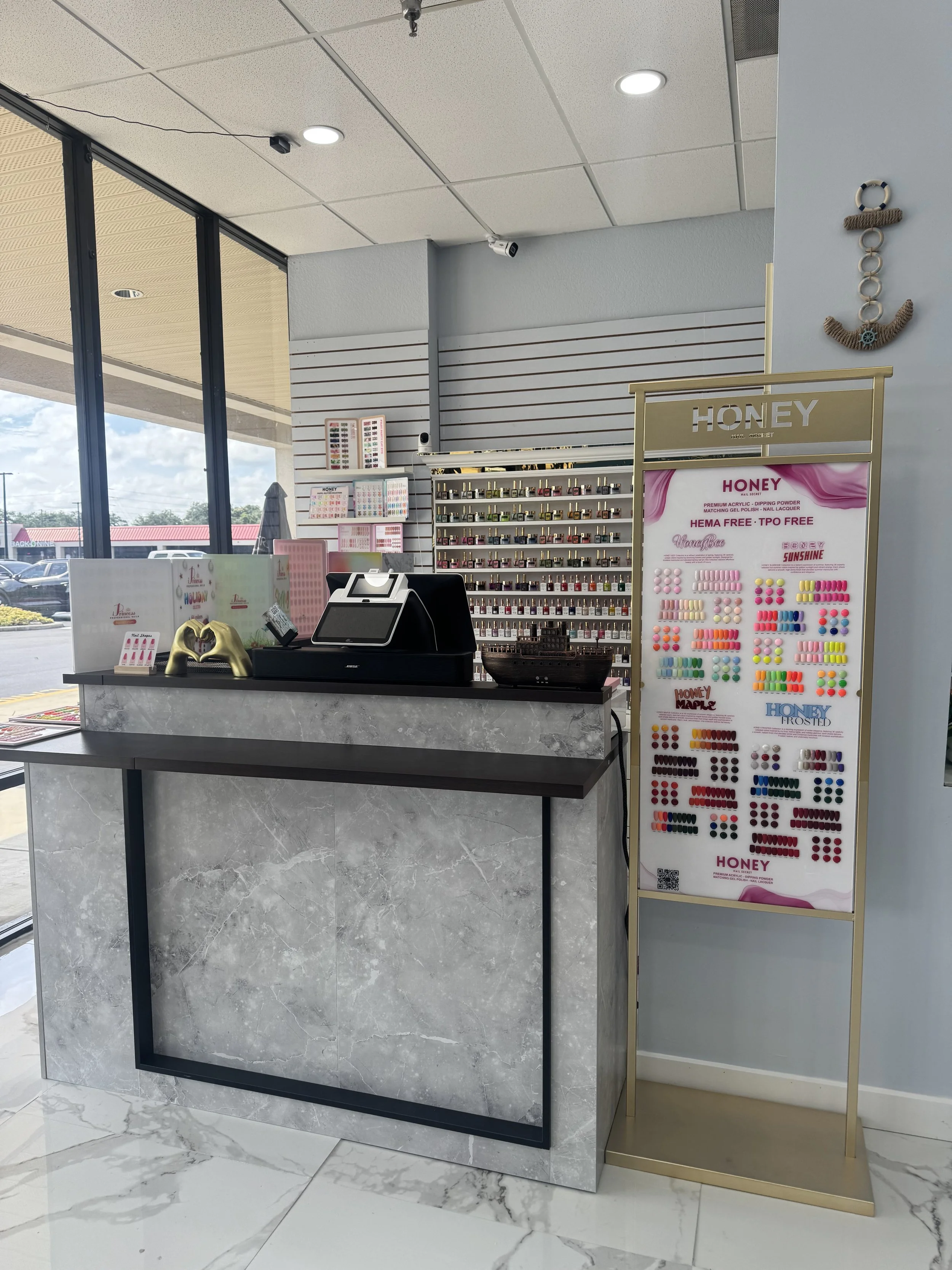 Inside a nail salon, a marble counter with a checkout register, a display of nail polish colors, and a stand with a chart of different nail polish options.