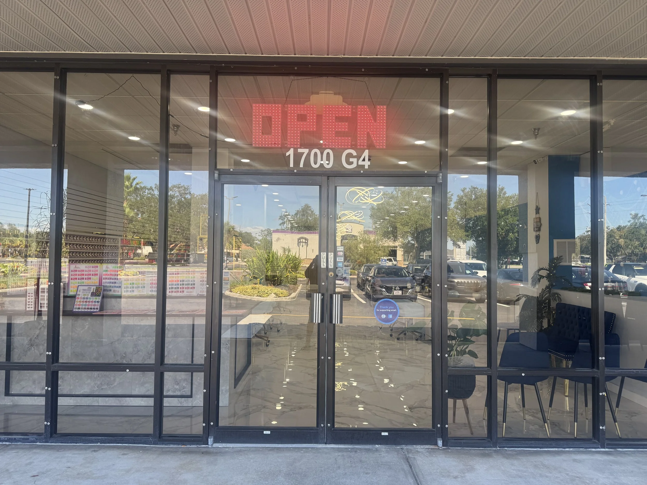 Store entrance with glass doors and a red LED 'OPEN' sign, located at 1700 G4. Inside the store, visible through the glass, there are chairs, tables, and decorative lights, with a parking lot outside visible through the windows.