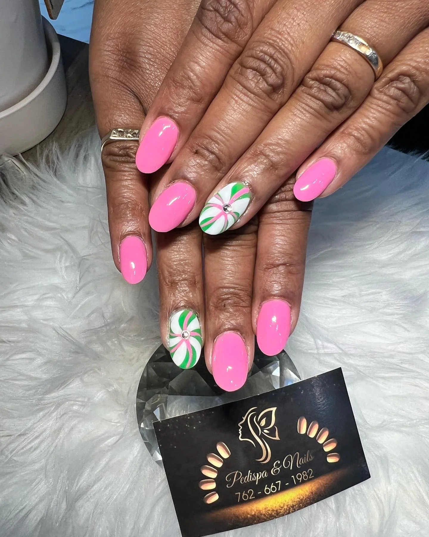Close-up of a person's hands with pink and green holiday-themed nail art, featuring peppermint swirl design and small rhinestone accents. The hands rest on a fluffy white surface, and a black business card for Pedispa & Nails is partially visible.