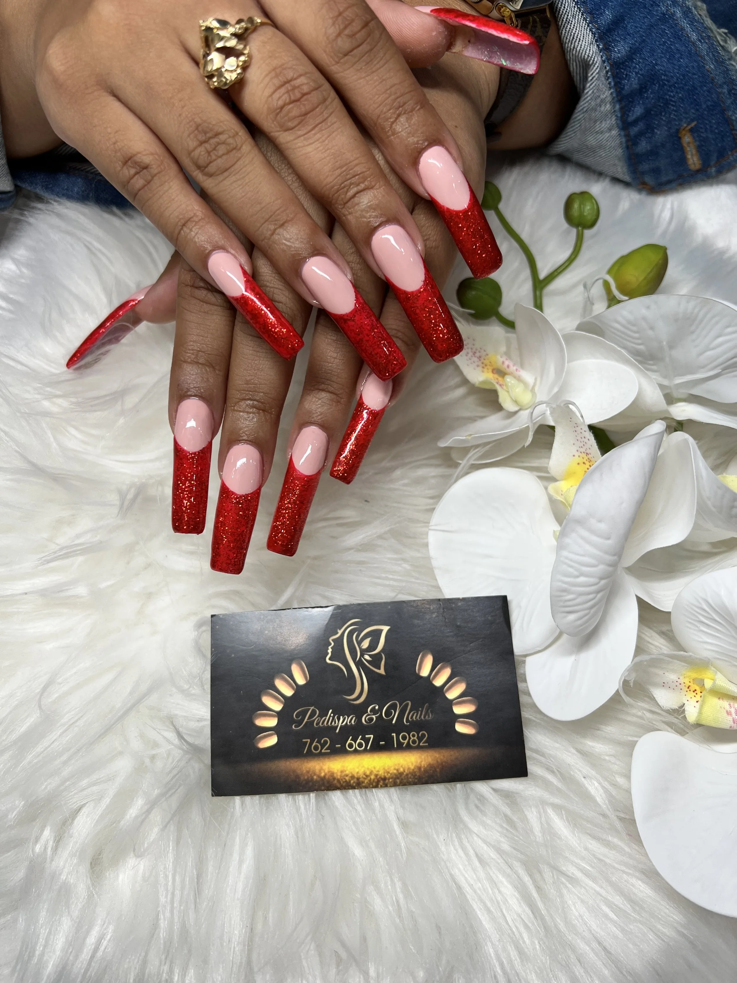 Close-up of a person's hand with long red and pink acrylic nails resting on a white fluffy surface, surrounded by white flowers. The person is wearing a gold ring and a denim jacket. In the foreground, there is a black business card for Pedispa & Nai