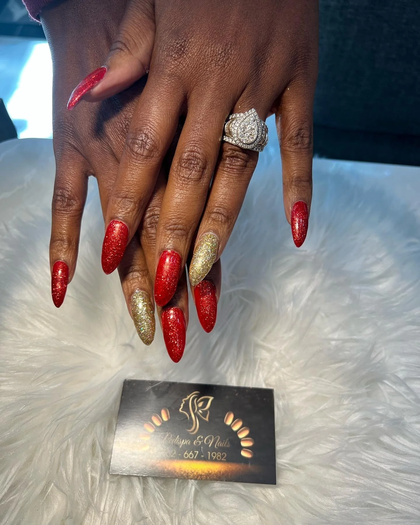 Close-up of a person's hands featuring long, almond-shaped red and gold glitter acrylic nails, with a large, ornate diamond engagement ring on the ring finger of the left hand, resting on a white marble surface.