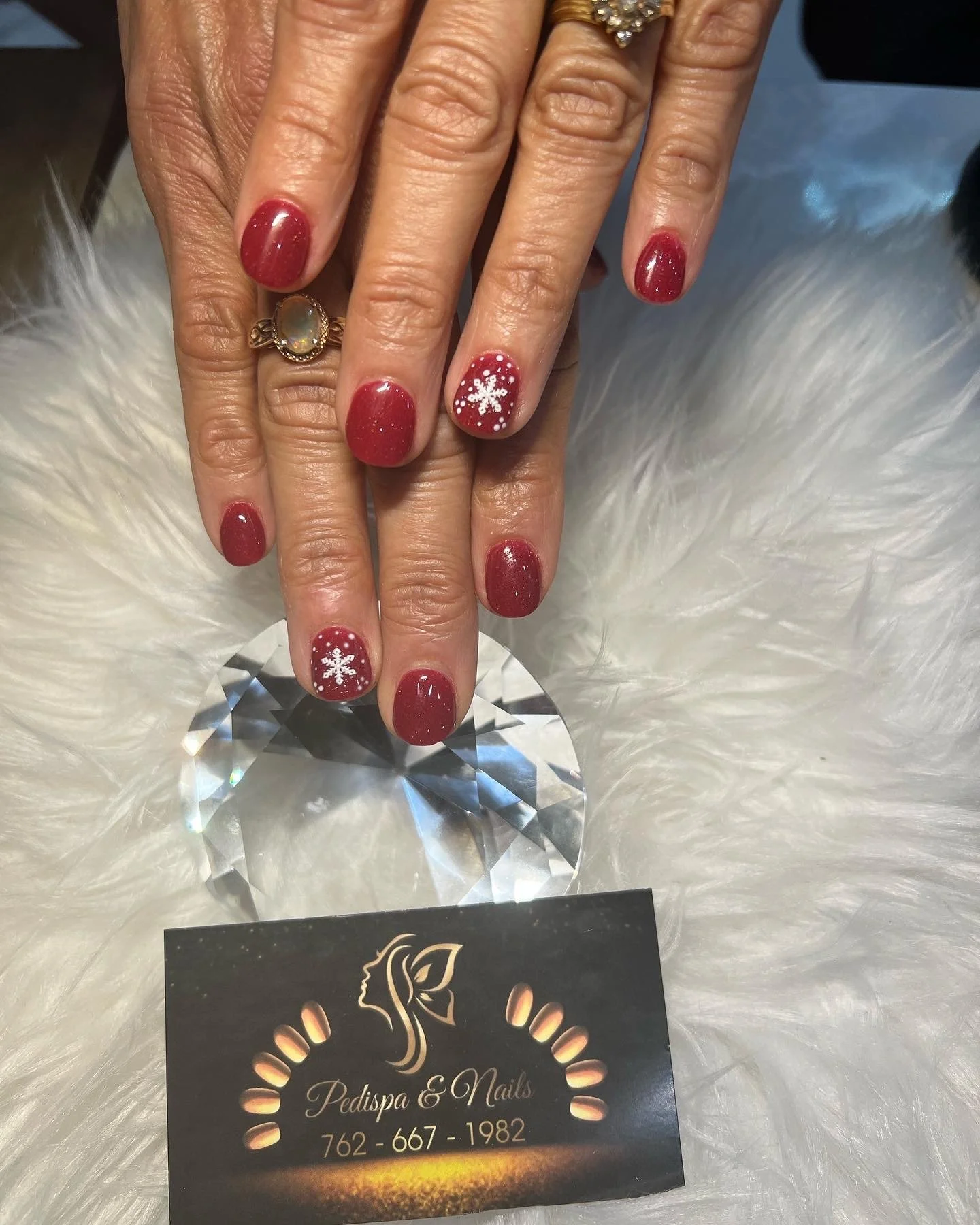 Close-up of a person's hands with manicured nails painted in red with tiny white snowflakes, on a white faux fur surface, with a large clear gemstone in the background and a black business card for Pedispa & Nails below.