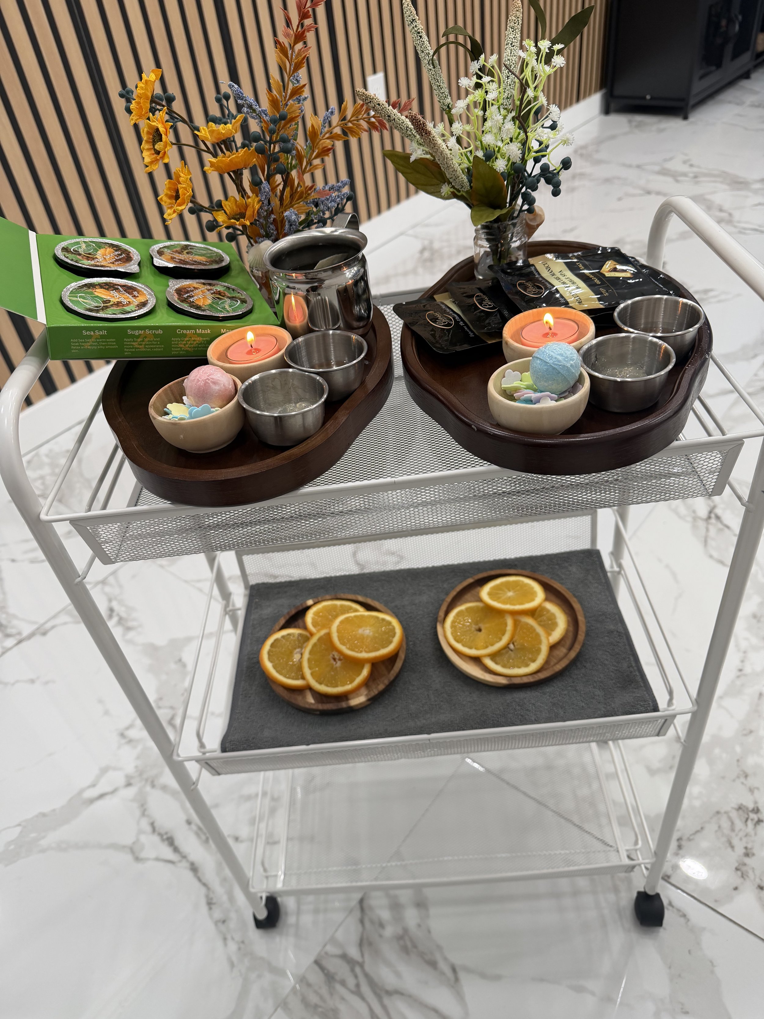 A white mobile cart with two shelves holding decorative items. The top shelf has two wooden trays with candles, small bowls, a potted plant with yellow and purple flowers, a metallic cup, and packets. The lower shelf holds a black tray with dried orange slices. The background features hardwood paneling and white marble floors.