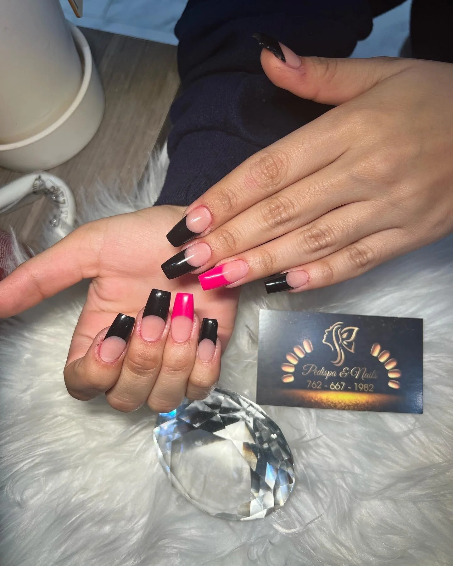 Close-up of a person's hand receiving a fresh manicure with black and pink acrylic nails at Pedispa & Nails, with a business card and a large diamond-shaped crystal on a fluffy white surface.