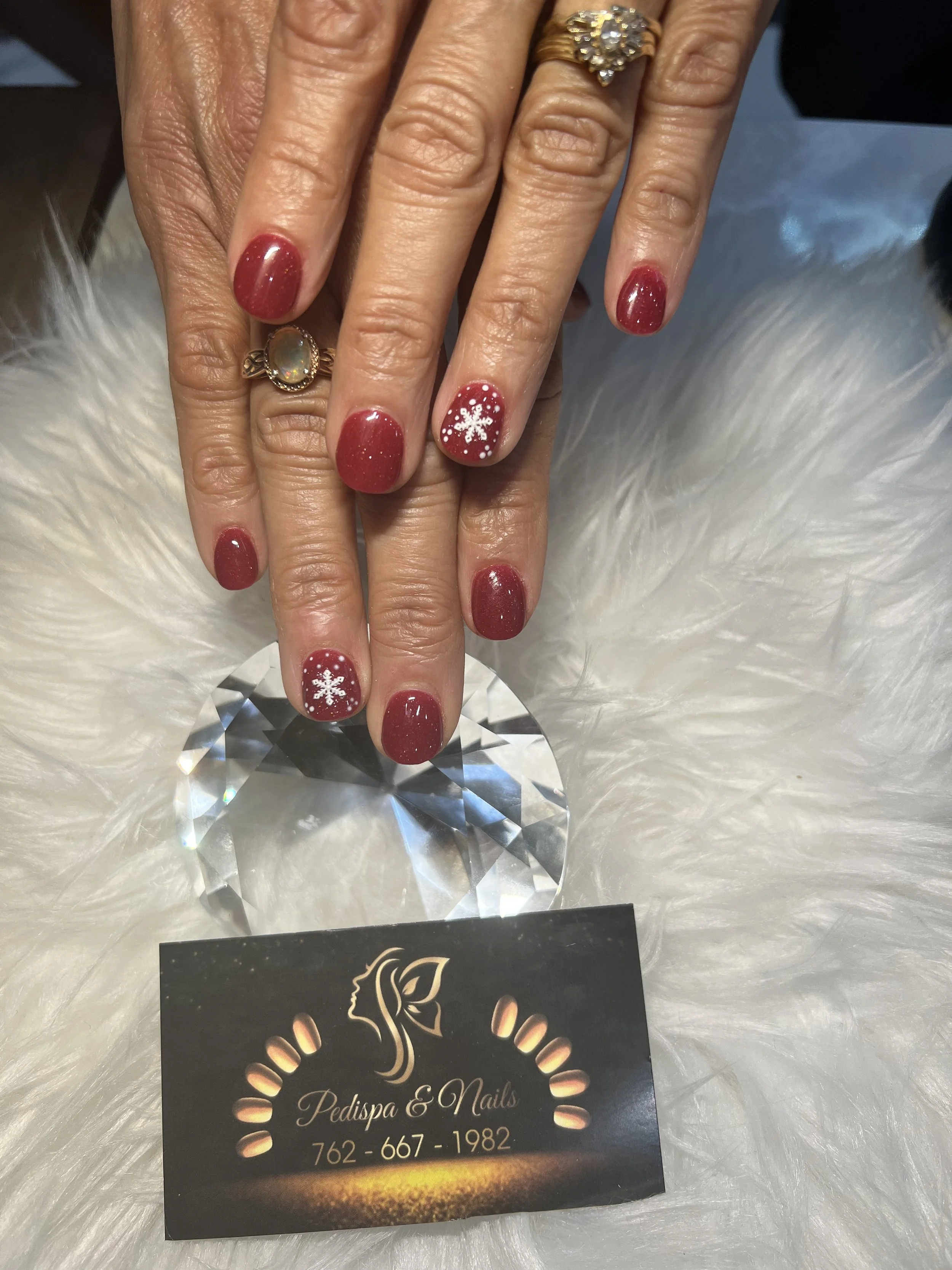 Hand with red glittery nails decorated with white snowflake design, wearing multiple rings, resting on a large, faceted crystal, with a business card for Pedispa & Nails partially visible beneath, set on a white, fluffy surface.