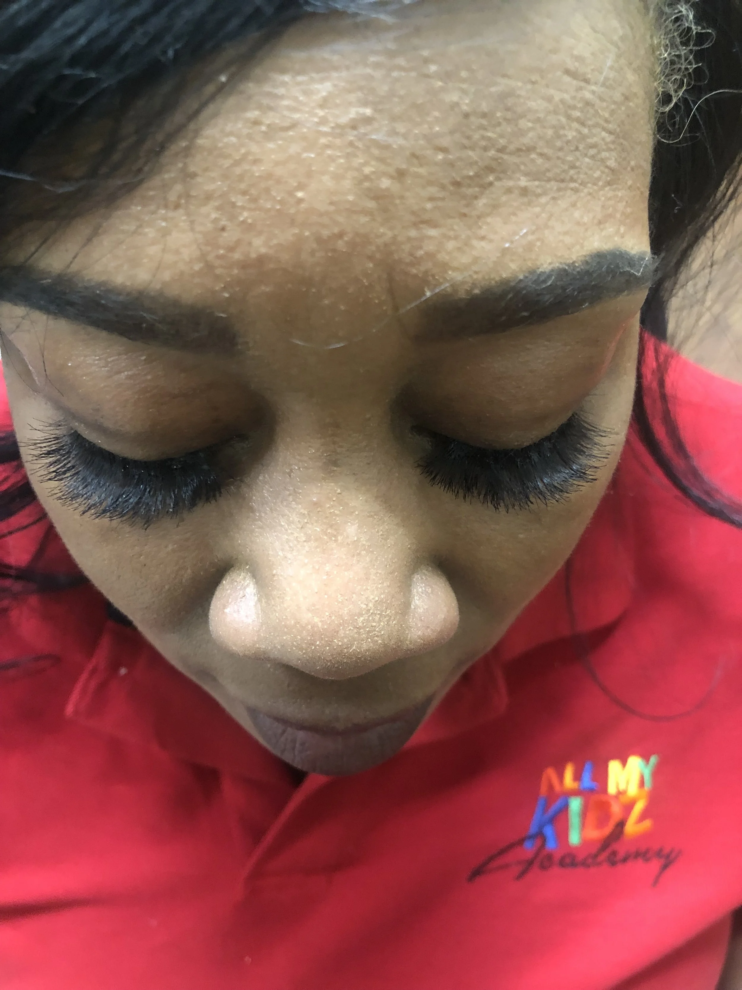 Close-up of a person's face from above, showing closed eyes, long eyelashes, and well-groomed eyebrows, with a red shirt visible in the lower part of the image.