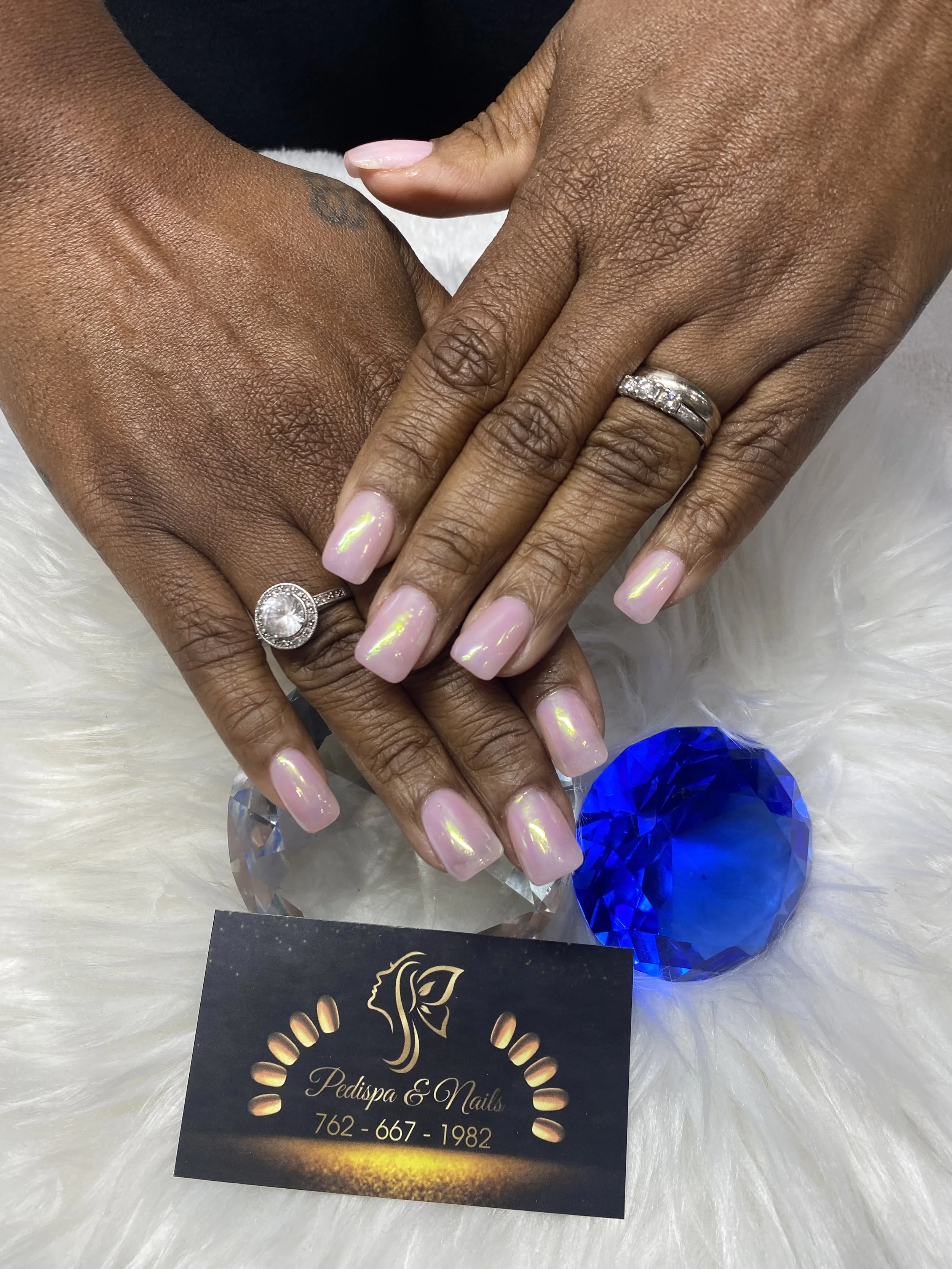 Close-up of two hands with manicured nails, one wearing a diamond ring, resting on a white furry surface, with a black business card and blue crystal decoration nearby.