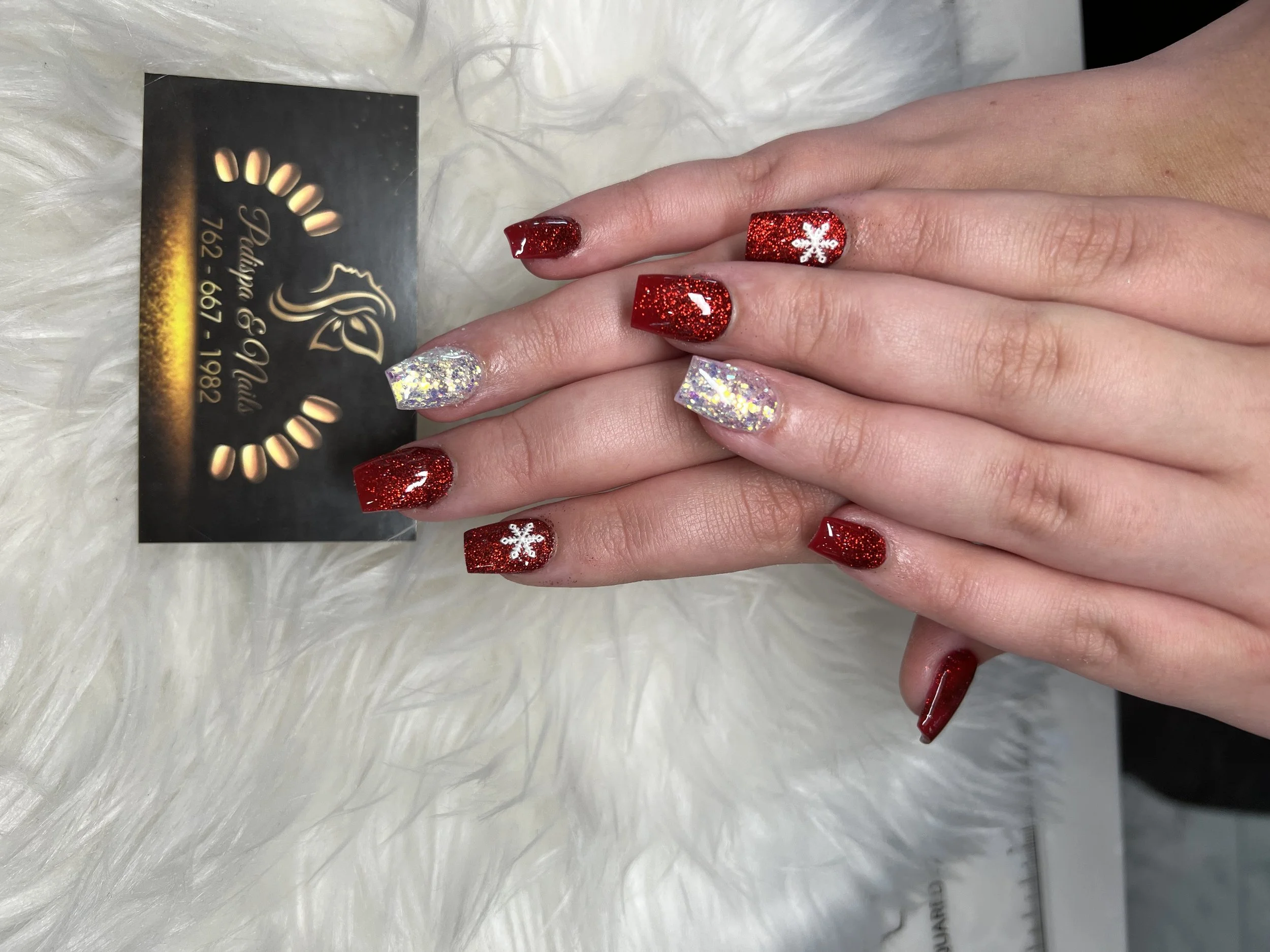 Close-up of hands with Christmas-themed nail art, including red glitter nails and one with a white snowflake, resting on a white fluffy surface. A black business card with gold and white text and logo is partially visible.