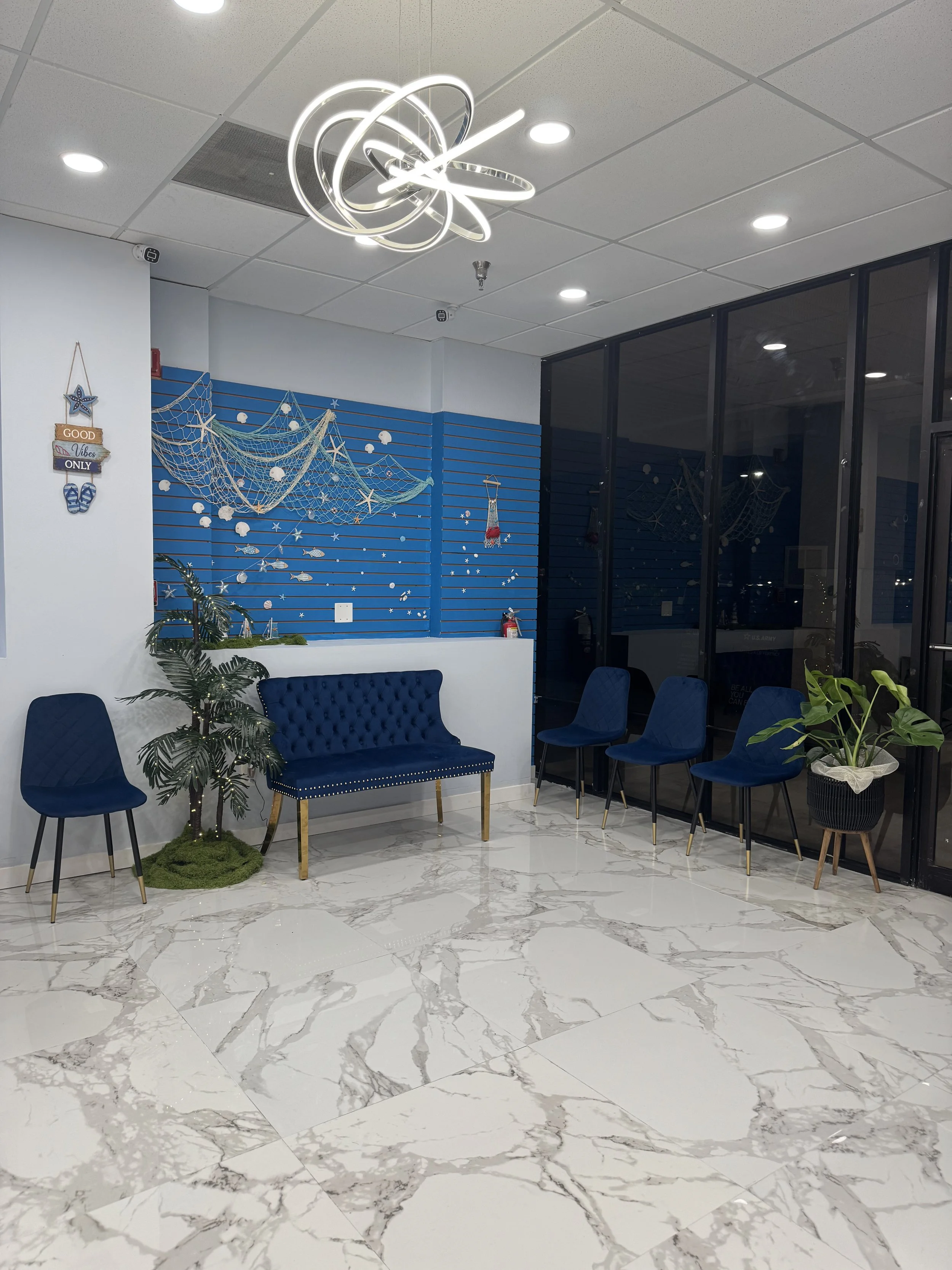 Interior space with white marble floor with gray veins, blue wall with nautical decorations including fishing nets, shells, and starfish, black framed glass wall, white ceiling with recessed lighting and modern chandelier, blue velvet chairs, green p