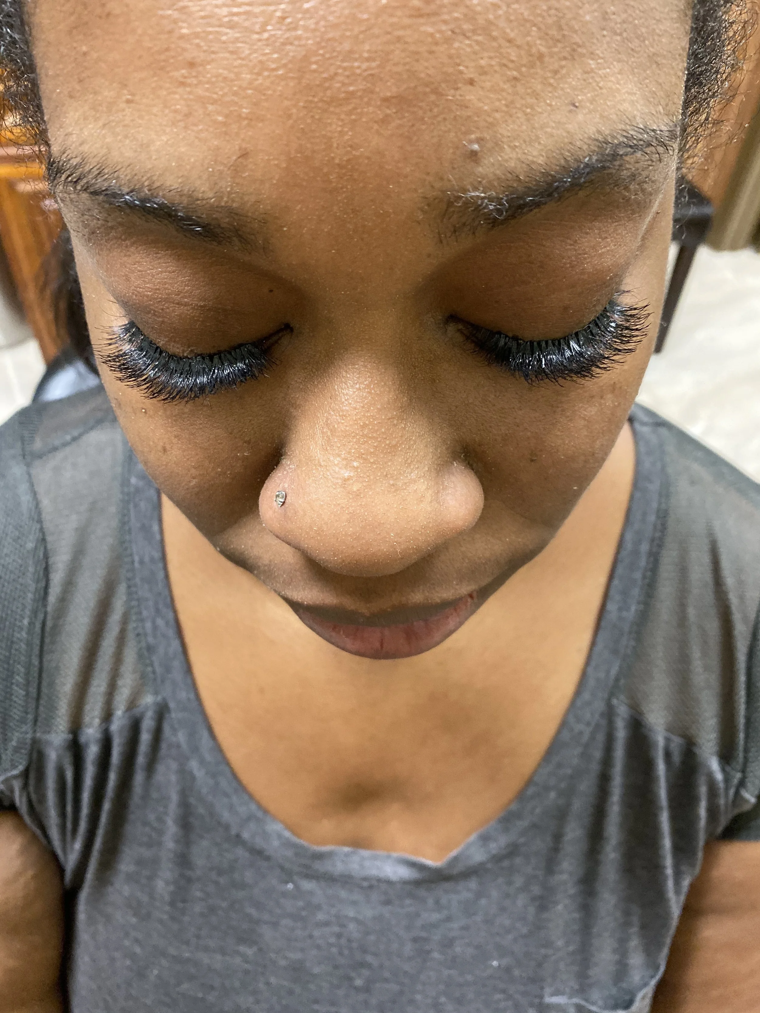 Close-up of a person's face looking downward, with closed eyes, dark skin, long eyelashes, and a small nose piercing.