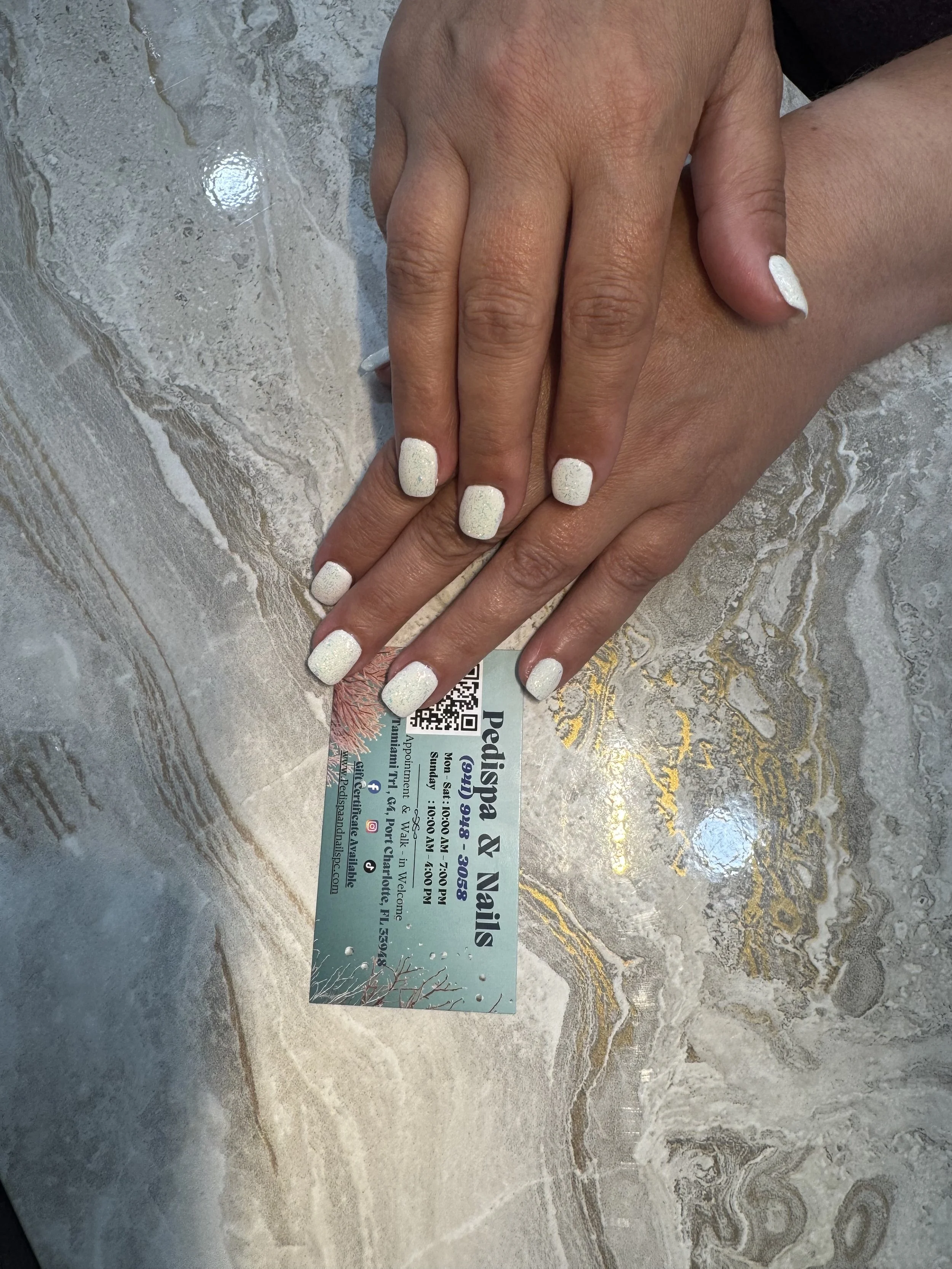 A pair of hands with white painted nails resting on a marble surface, with a business card partially visible underneath.