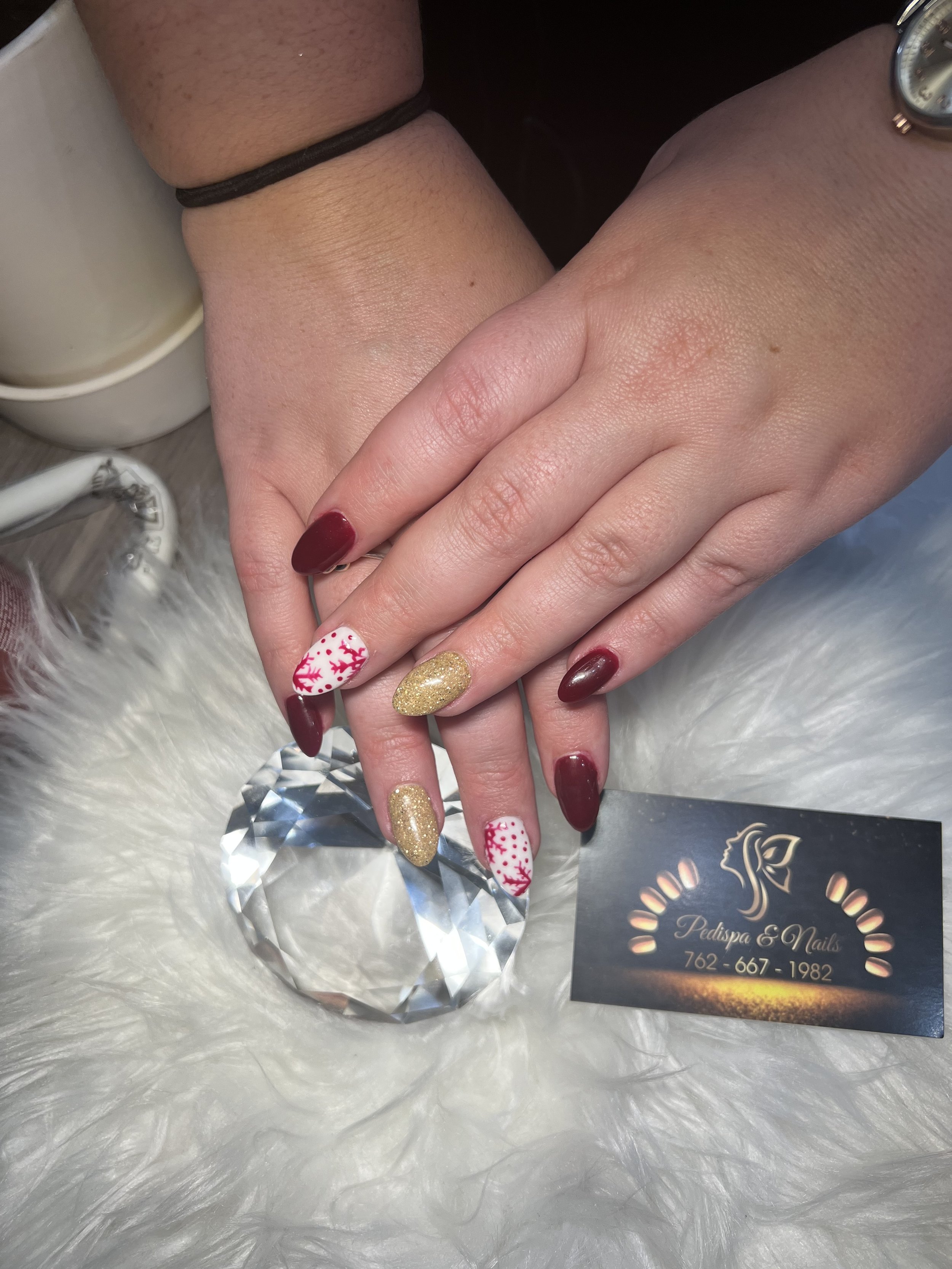 Close-up of a person's hand with decorated nails, resting on a fluffy white surface. The nails are painted in red, gold, and white with a Christmas theme. There is a business card for Pedispa & Nails under the hand, along with a large clear gem.