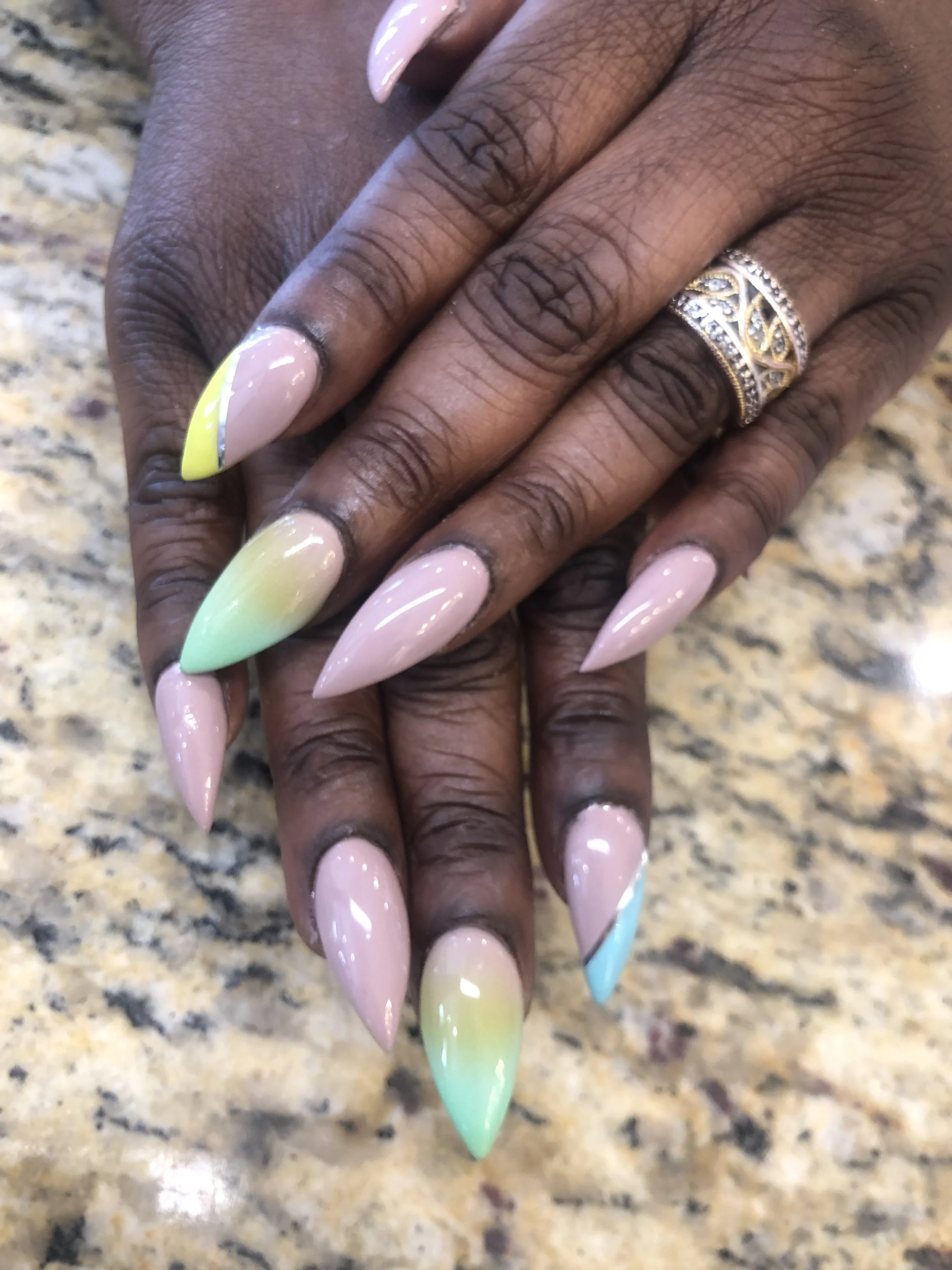 Close-up of a hand with long, pointed, pastel-colored nails in yellow, pink, green, and blue gradient, wearing multiple silver rings, resting on a granite countertop.