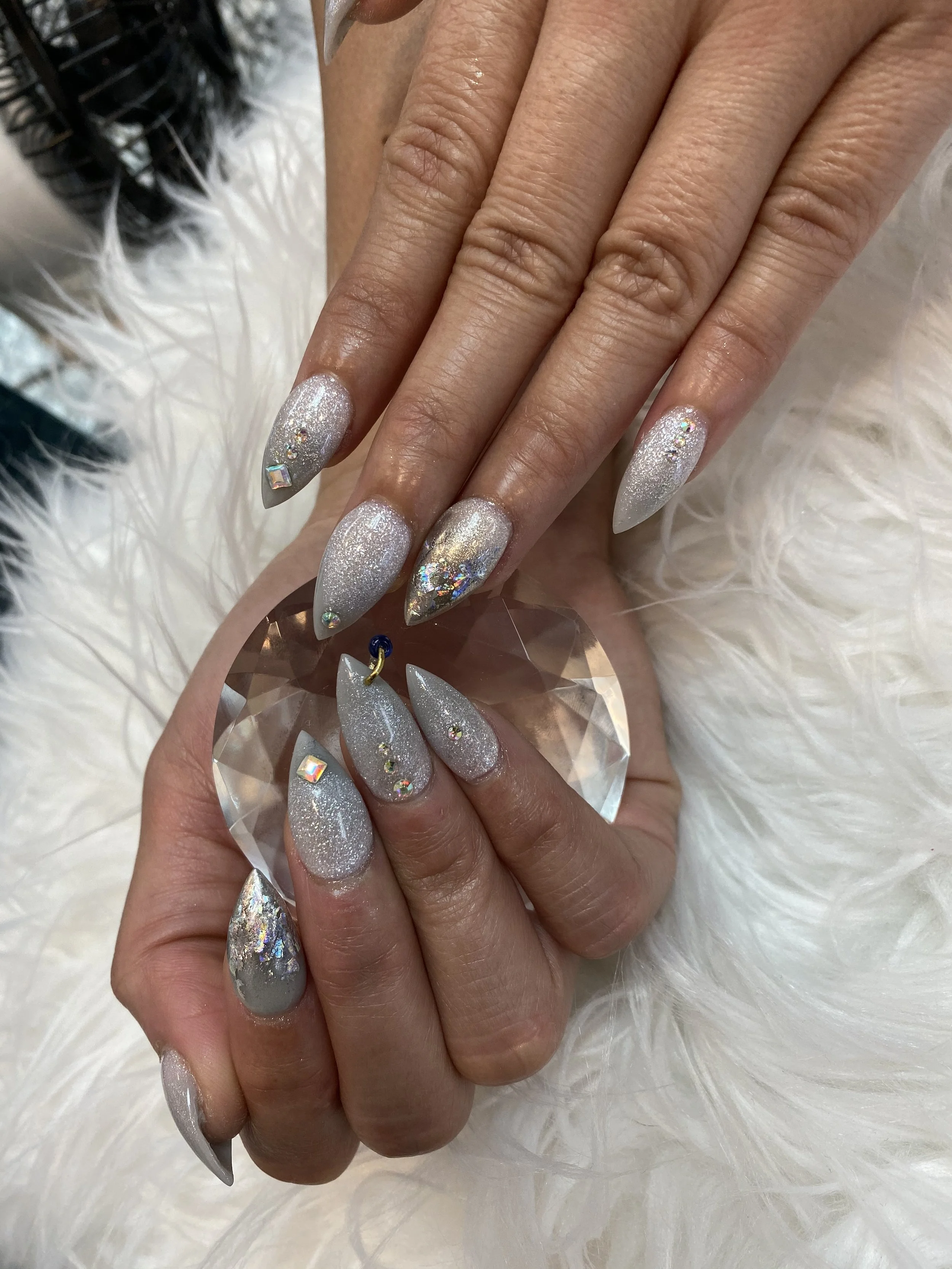 Close-up of a person's hand with long almond-shaped nails painted with a shimmery silver polish and decorated with small, multicolored rhinestones. The hand is resting on a soft, fluffy white surface.