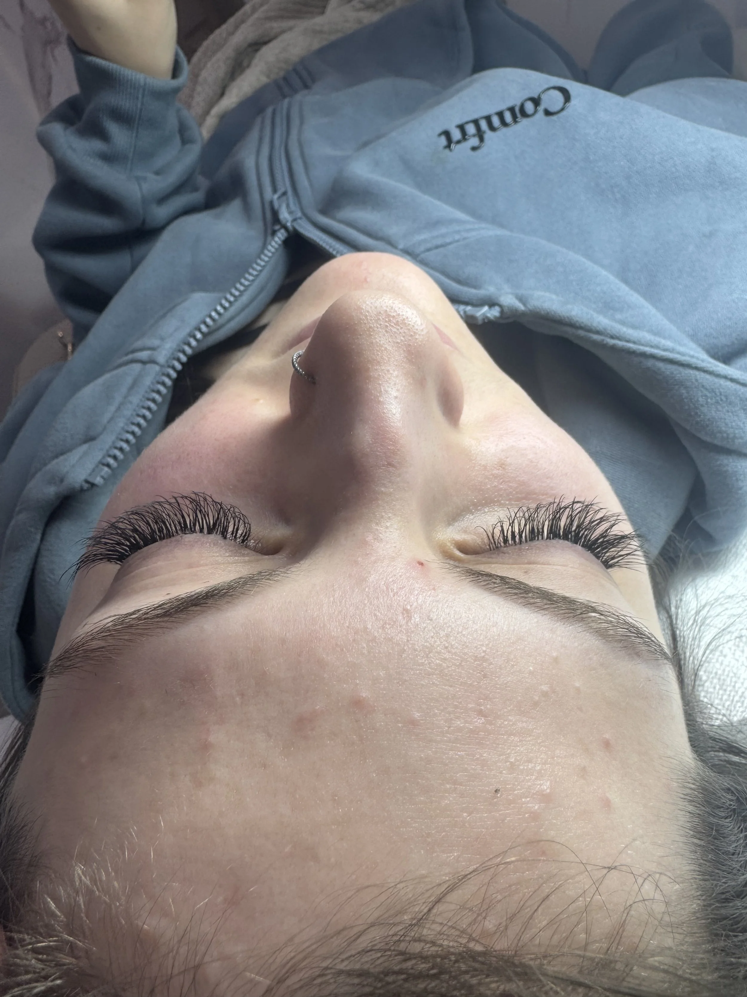 A person with long eyelashes lying down with their eyes closed, wearing a gray and blue hoodie with the word 'Comhf' visible, with a nose ring and some facial blemishes.