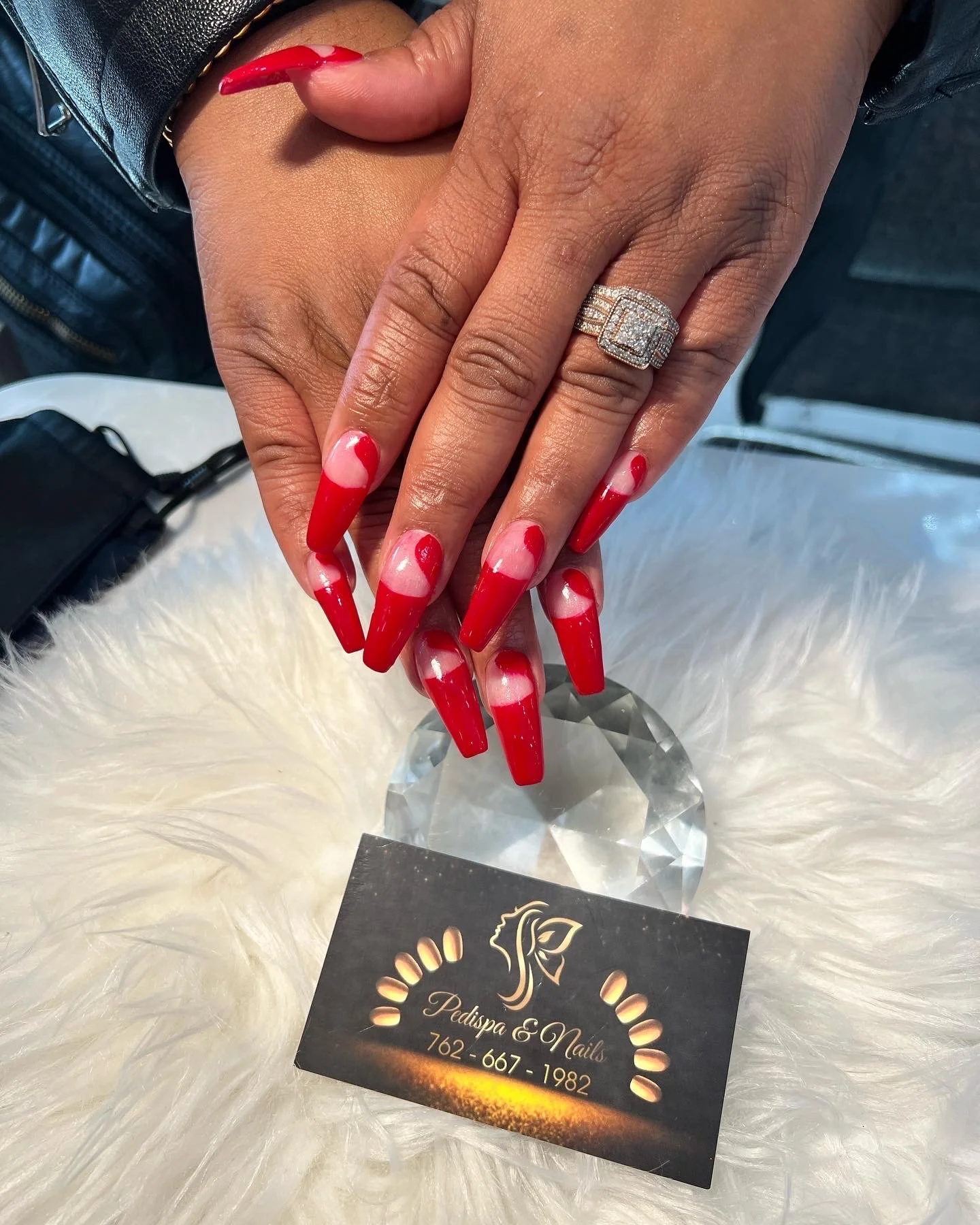 Hand with long red and pink acrylic nails, wearing a large diamond ring, resting on a fluffy white surface. There is a business card for Pedipa & Nails with a phone number and logo visible nearby.