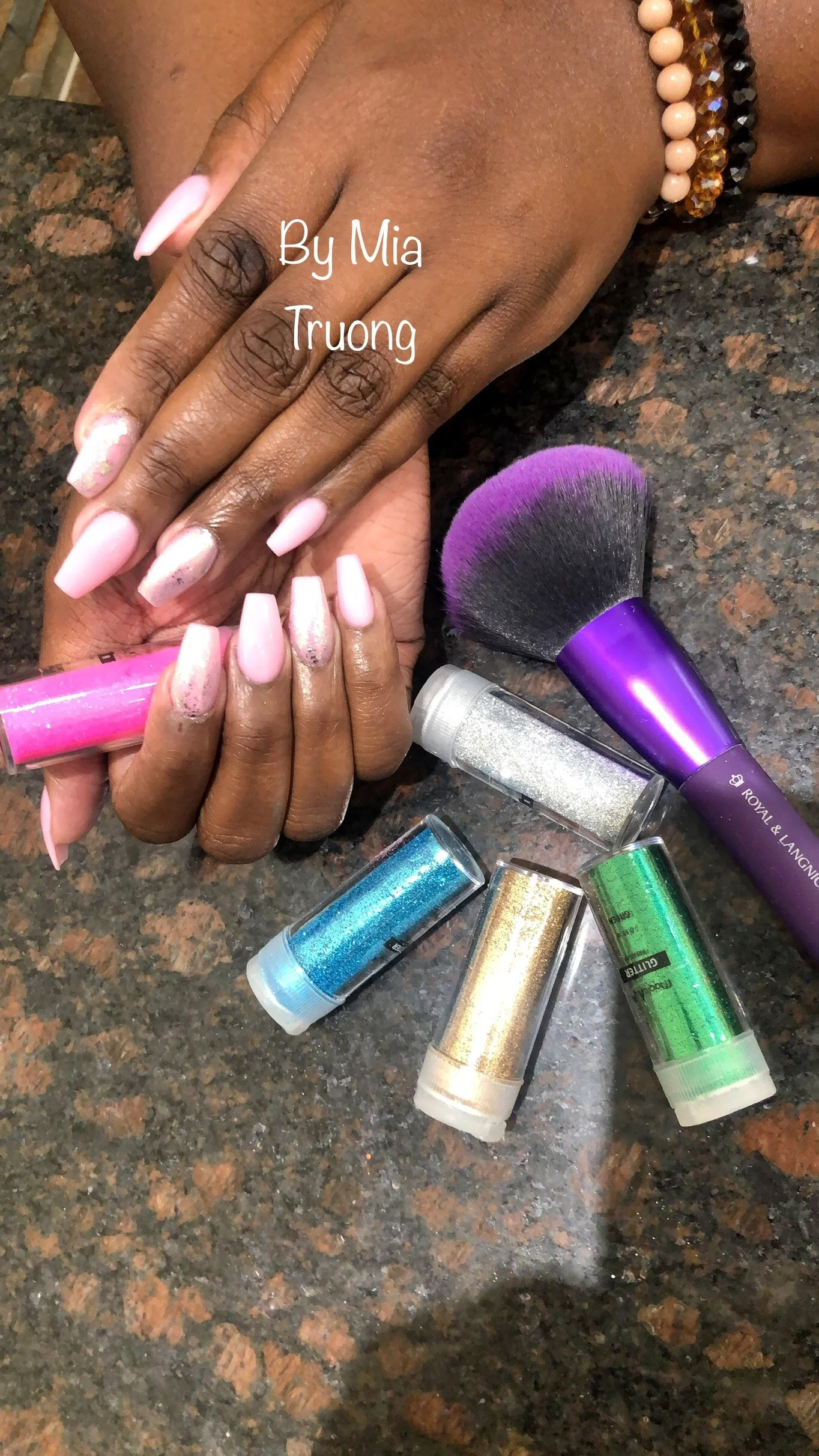 A person's hand with light pink manicured nails resting on a dark granite countertop surrounded by glitter bottles, a makeup brush, and a hand with darker skin tone. The text 'By Mia Truong' is overlaid on the hand.