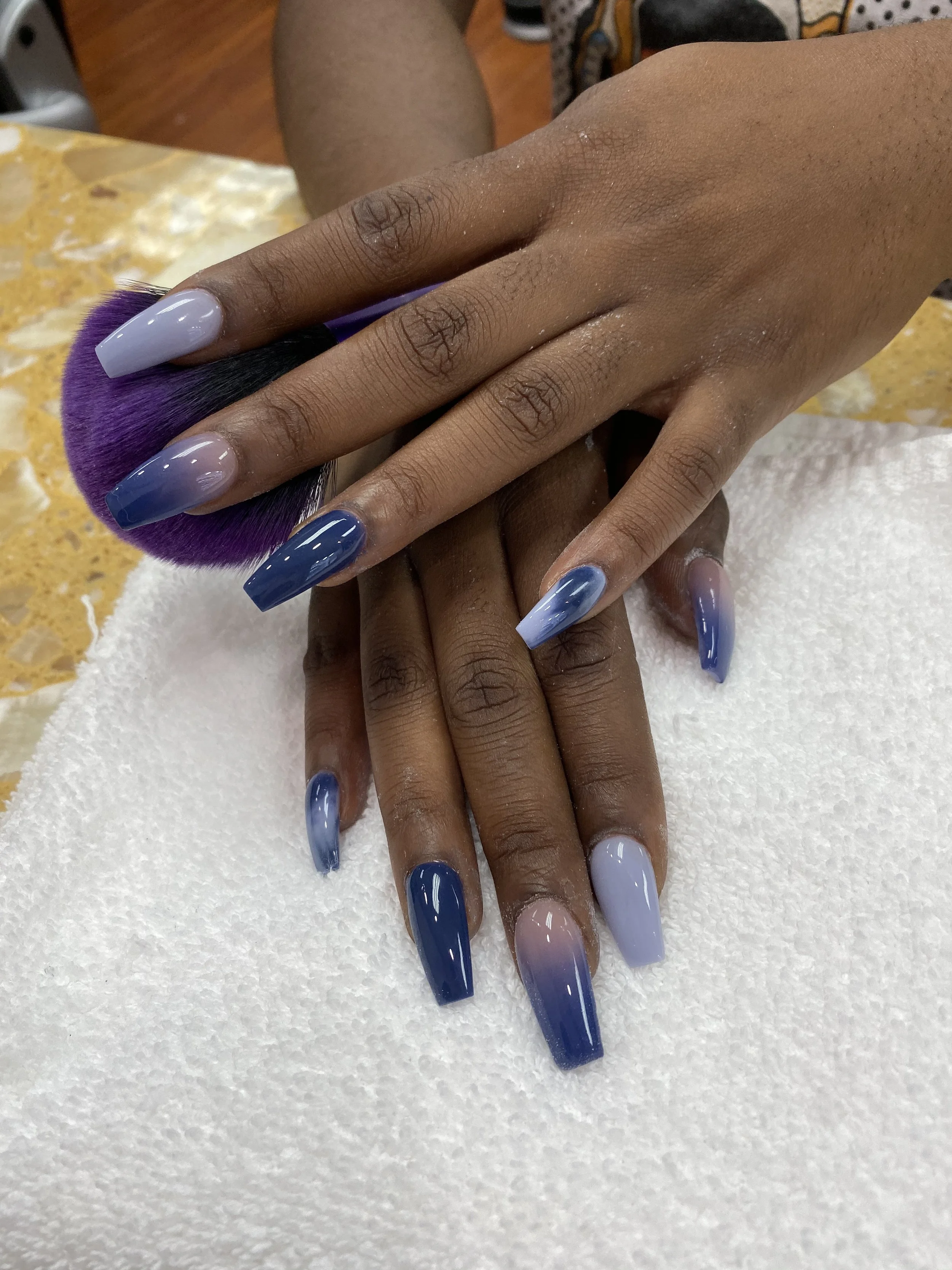 A person's hand with long, square-shaped nails painted in various shades of blue, resting on a purple makeup brush. The background includes a marble countertop and some shoes.