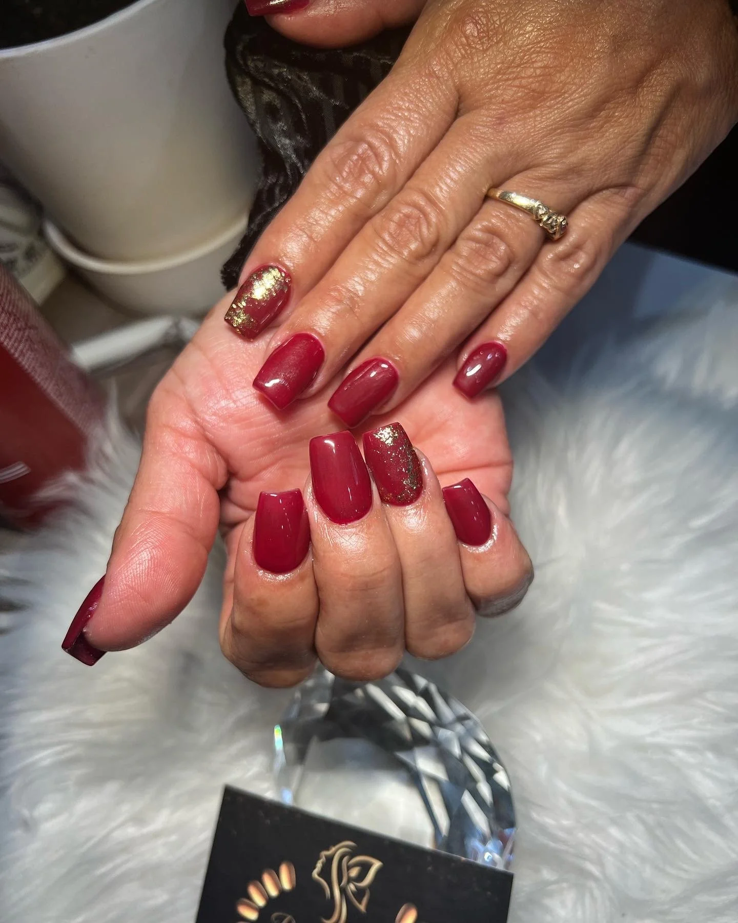 Close-up of a person's hand displaying freshly manicured nails painted in deep red with gold glitter accents, wearing a gold wedding band on the ring finger.