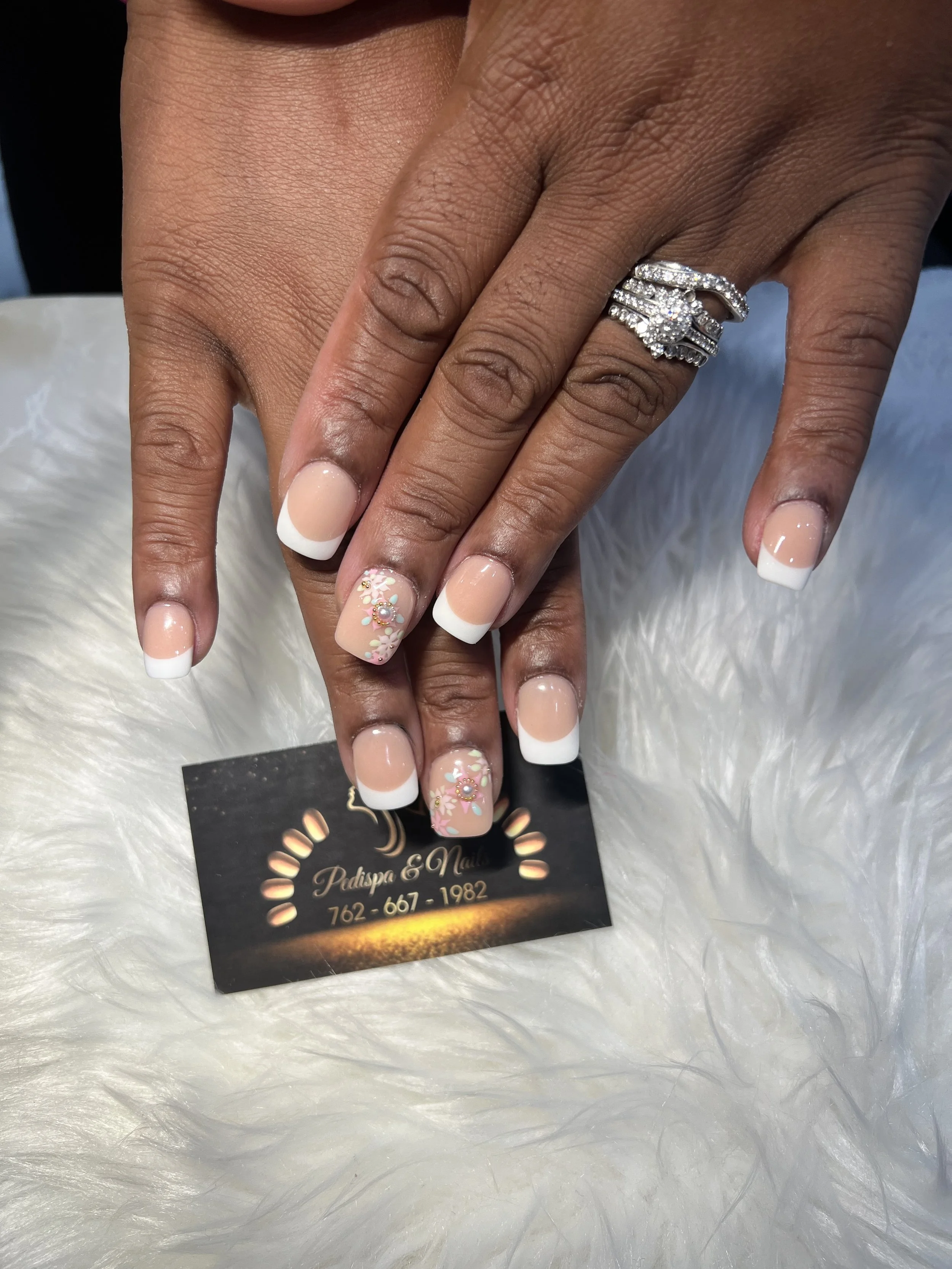 A person's hand with a ring, displaying a French manicure with floral accents, resting on a white fluffy surface. There is also a business card underneath the hand with the text 'Pedspa & Nails' and contact number.