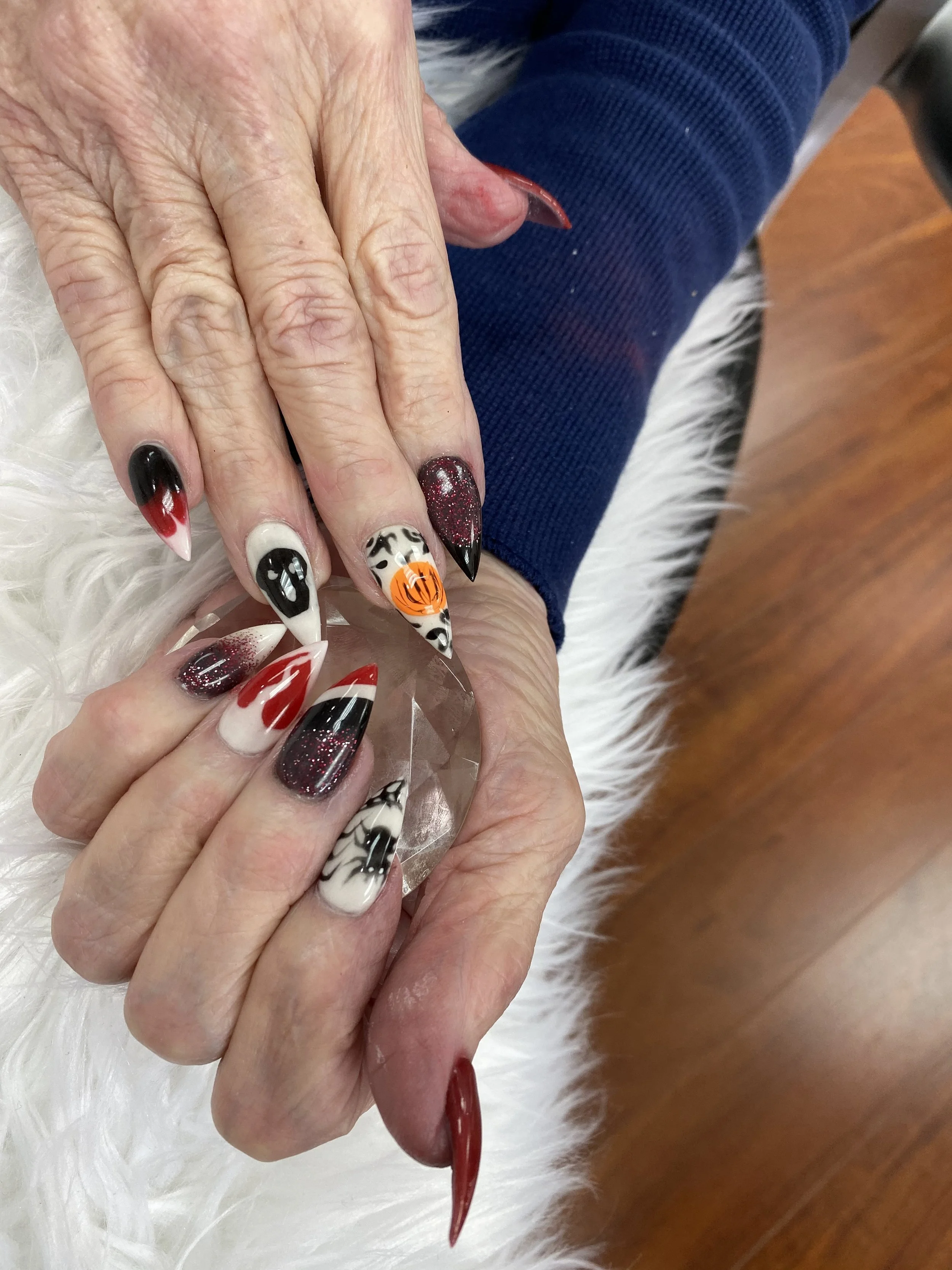 Close-up of an elderly person's hand with elaborately decorated Halloween-themed nail art, featuring black, white, red, and orange colors, resting on a white fluffy surface.