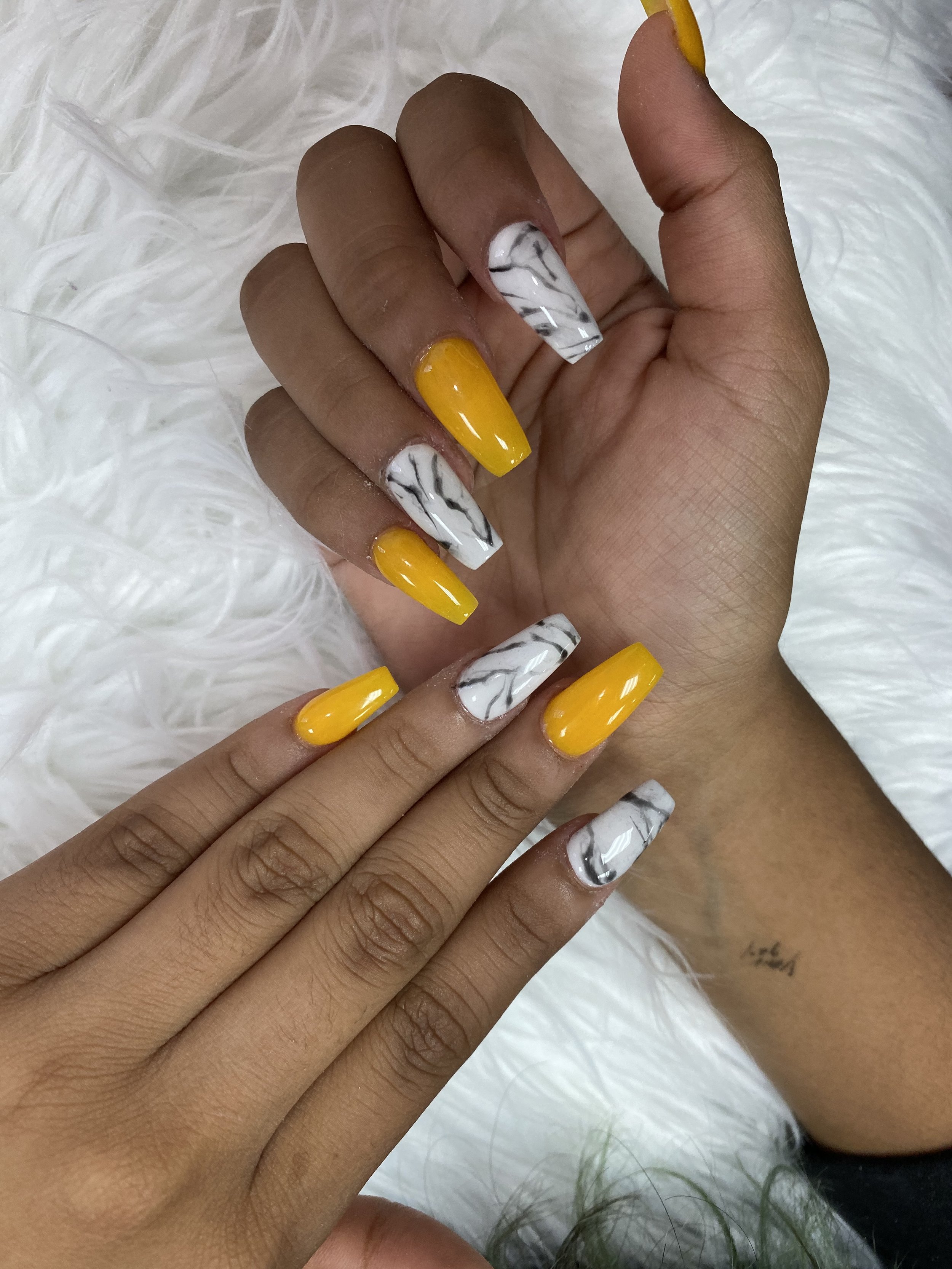 Close-up of a person's hand with yellow and white marble-effect painted nails, resting on a soft white fur background.