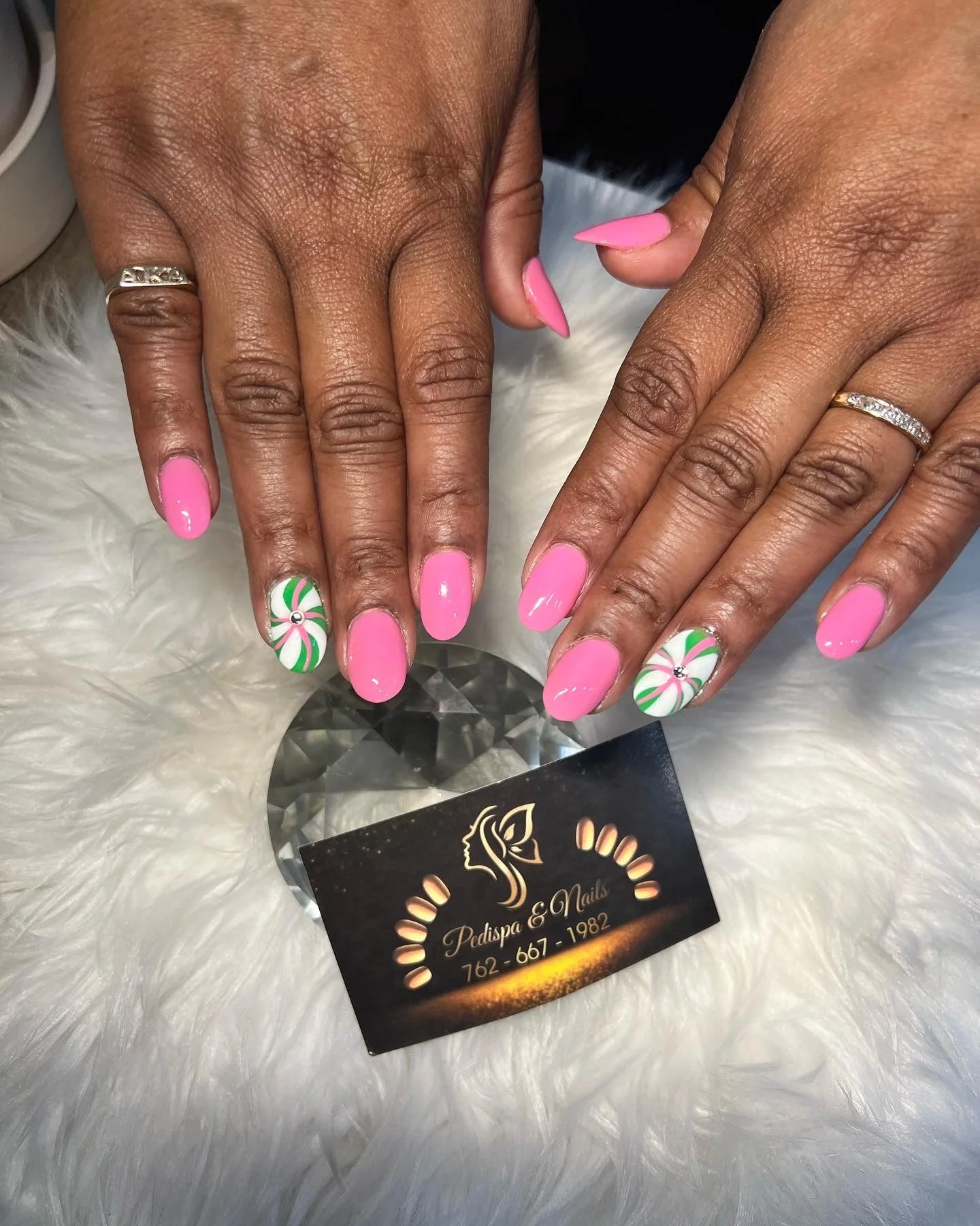 Close-up of two hands with pink manicure, one hand has Christmas-themed nail art with green and white swirl and a small gem. Both hands have rings, one with a gold band and embedded stones.