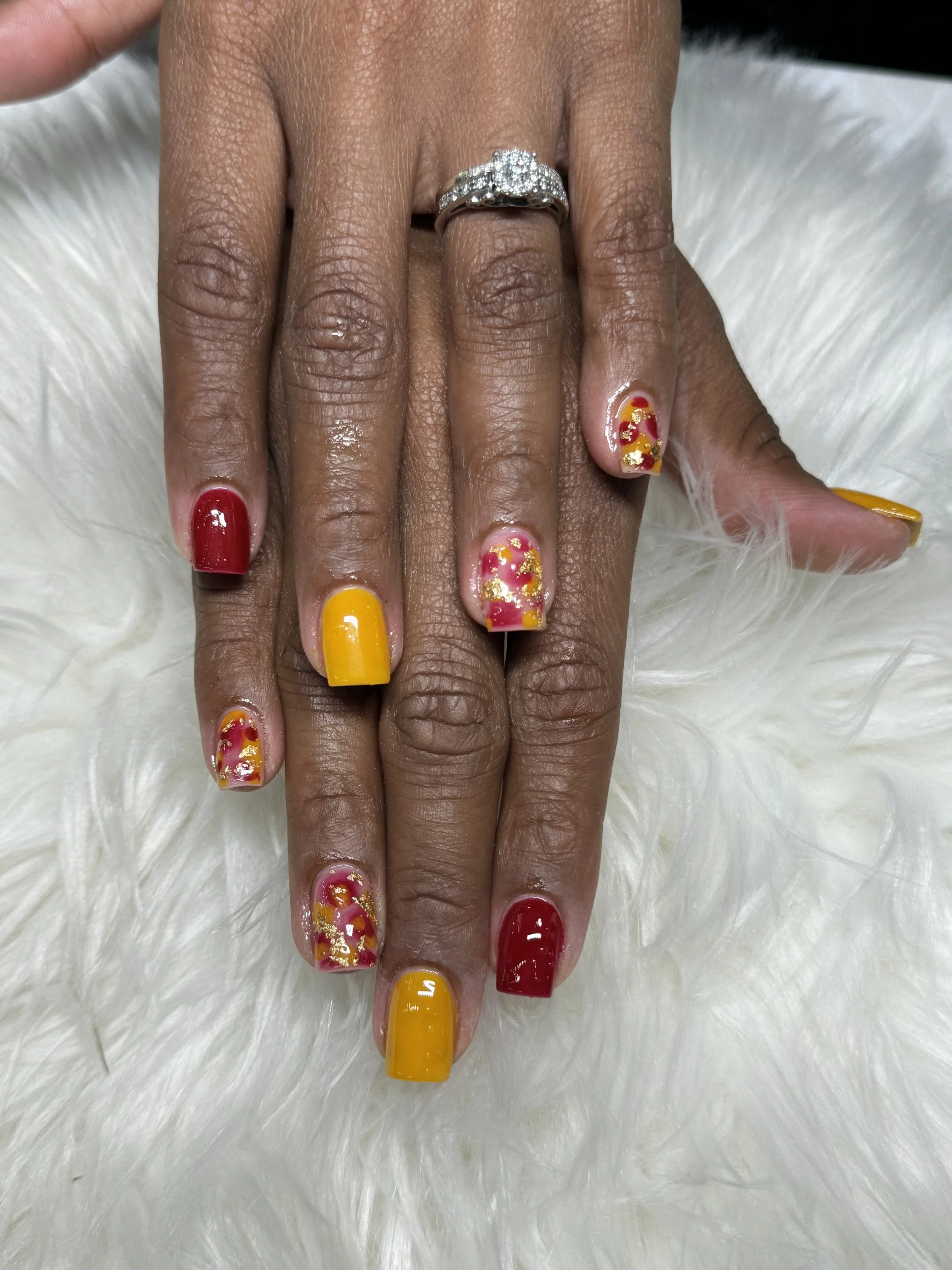 Hand with colorful nail polish featuring red, yellow, and floral designs, resting on a white furry surface.