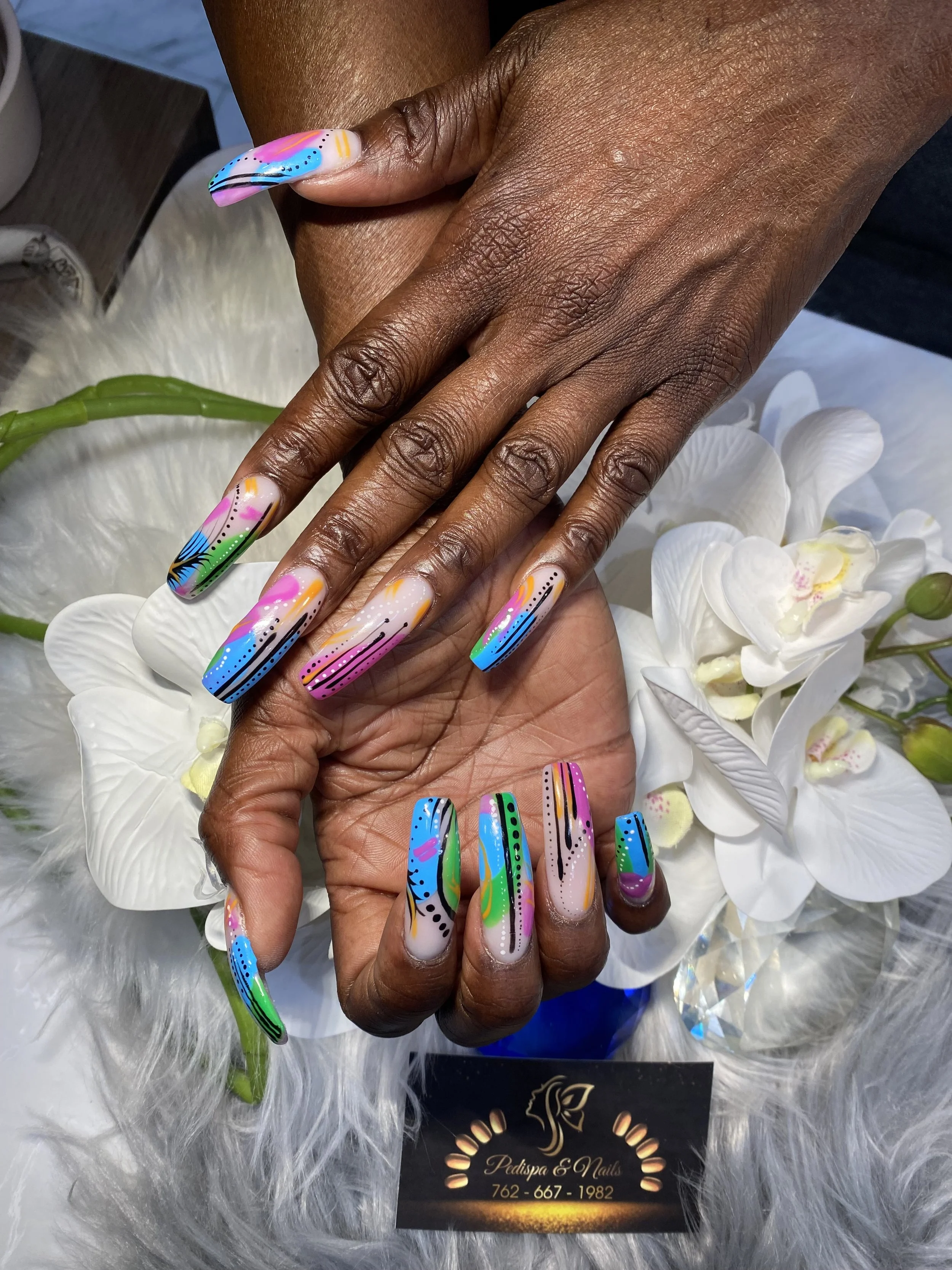 Close-up of a person's hand with colorful, abstract nail art, resting on a white floral arrangement and furry fabric, with a business card for Pedispa & Nails visible in the background.