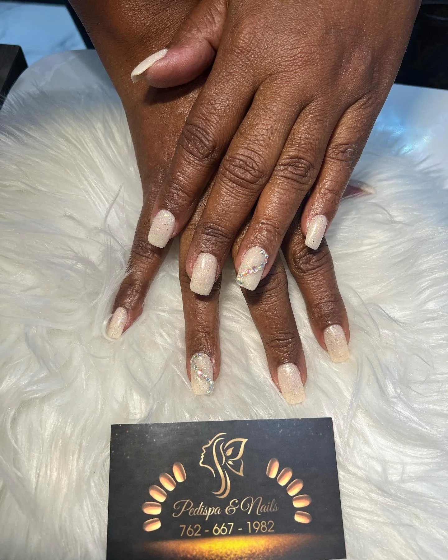 A pair of hands with manicured nails, featuring light-colored polish with glitter and rhinestone accents. The hands rest on a white fluffy surface, with a business card for Pedispa & Nails partially visible beneath them.