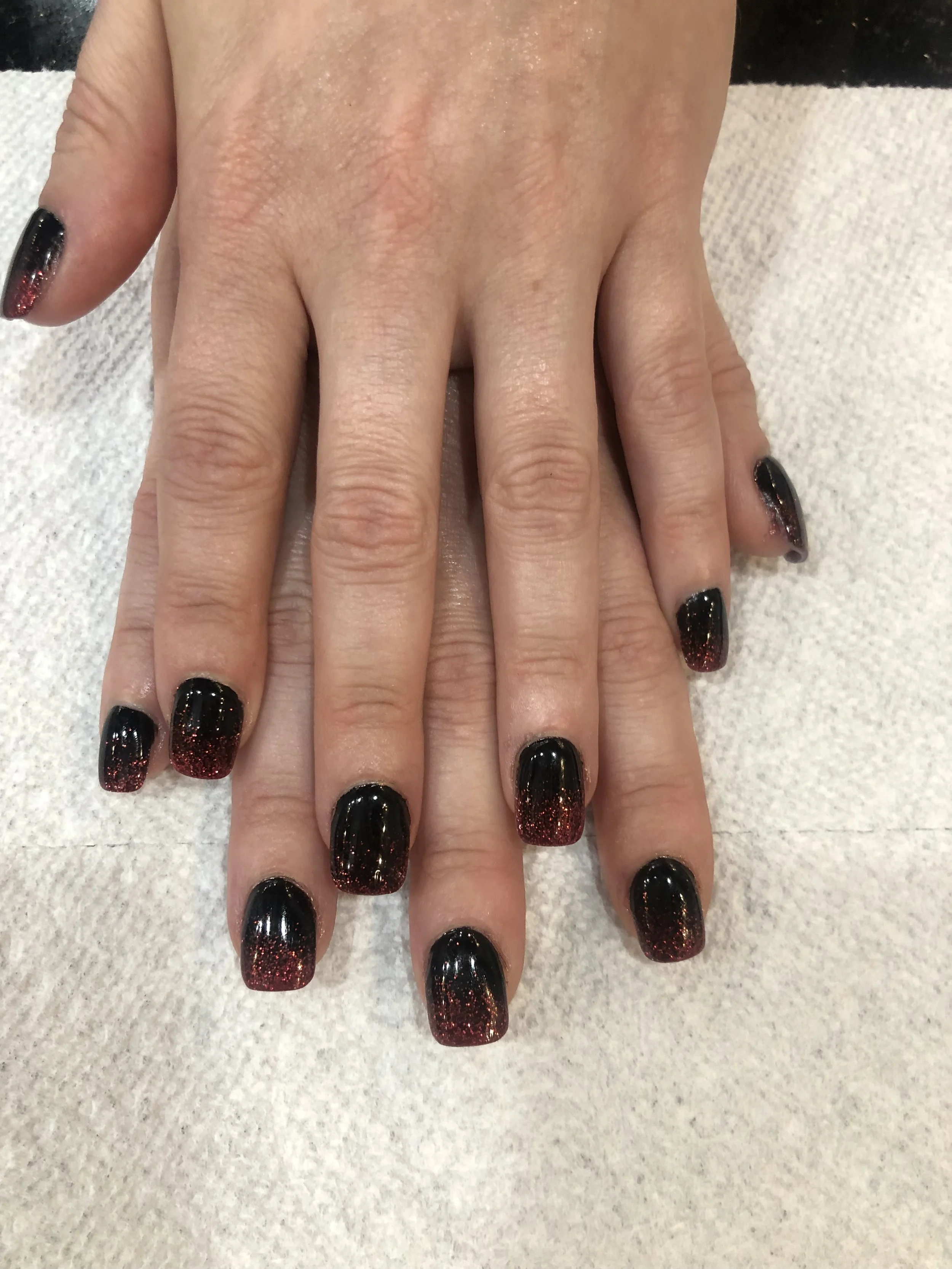 Close-up of two hands with black and red glitter nail polish, resting on a white textured cloth.