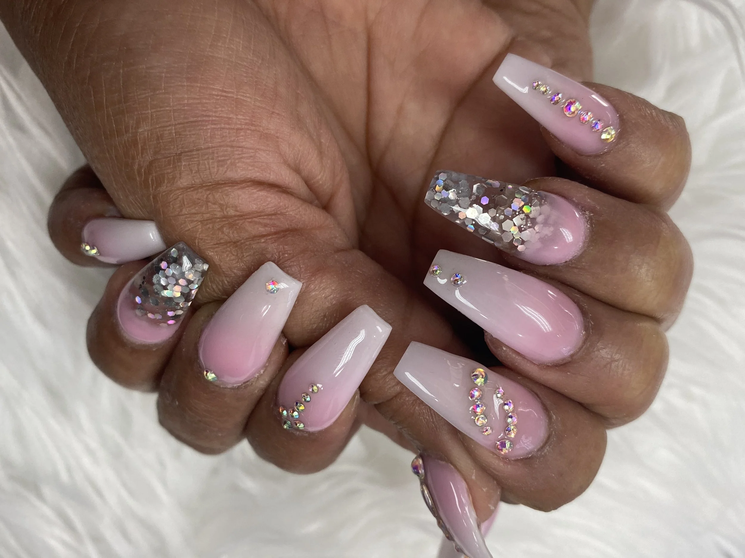 Close-up of a hand with long, square-shaped nails painted in a soft pink color. The nails are decorated with multicolored rhinestones and glitter accent nails, resembling a glamorous, sparkling style.
