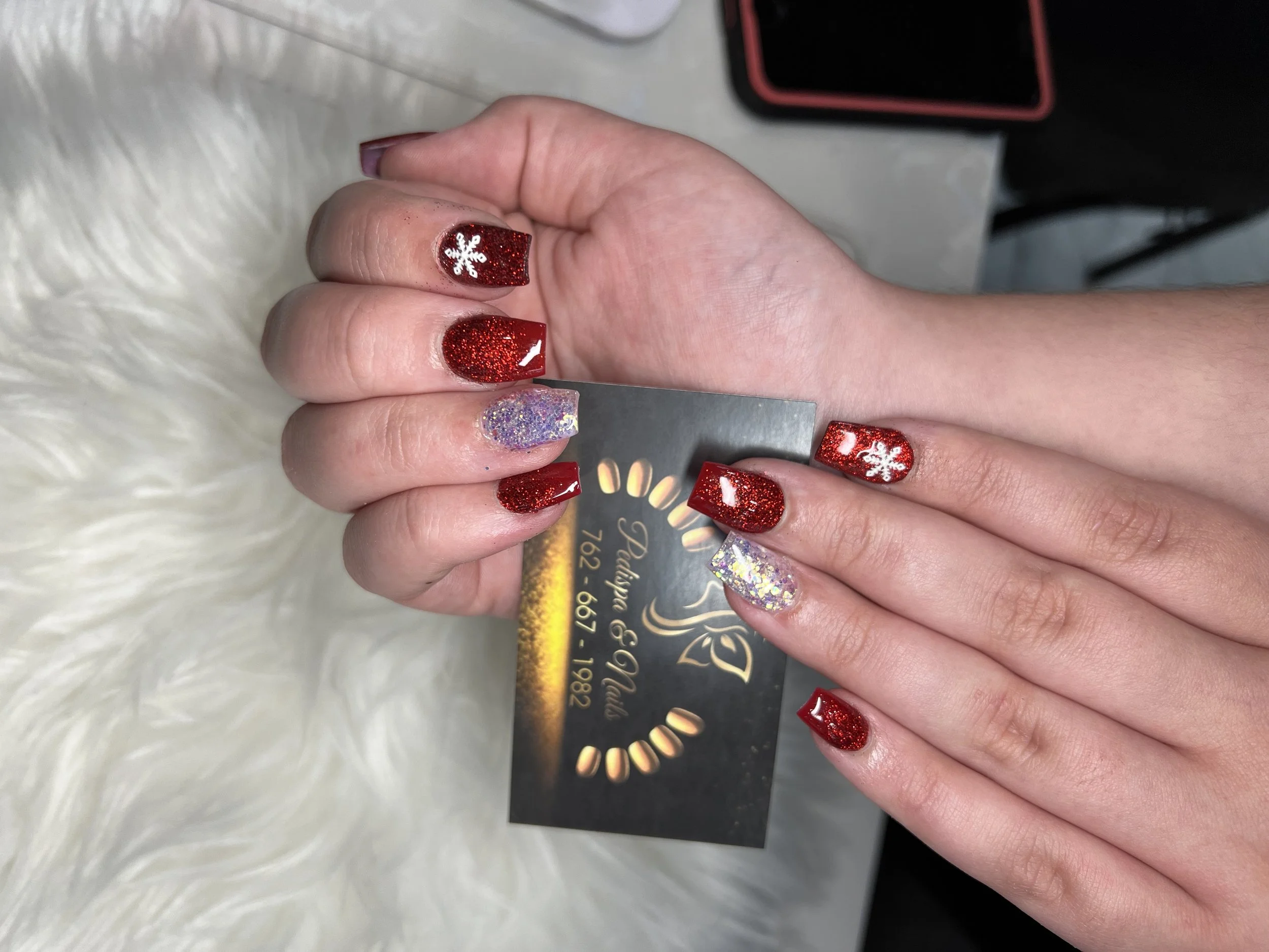 Close-up of a person's hand showing freshly manicured nails with red glitter nail polish and white snowflake designs on some nails. The hand is resting on a white furry surface, and there are black and red objects in the background.