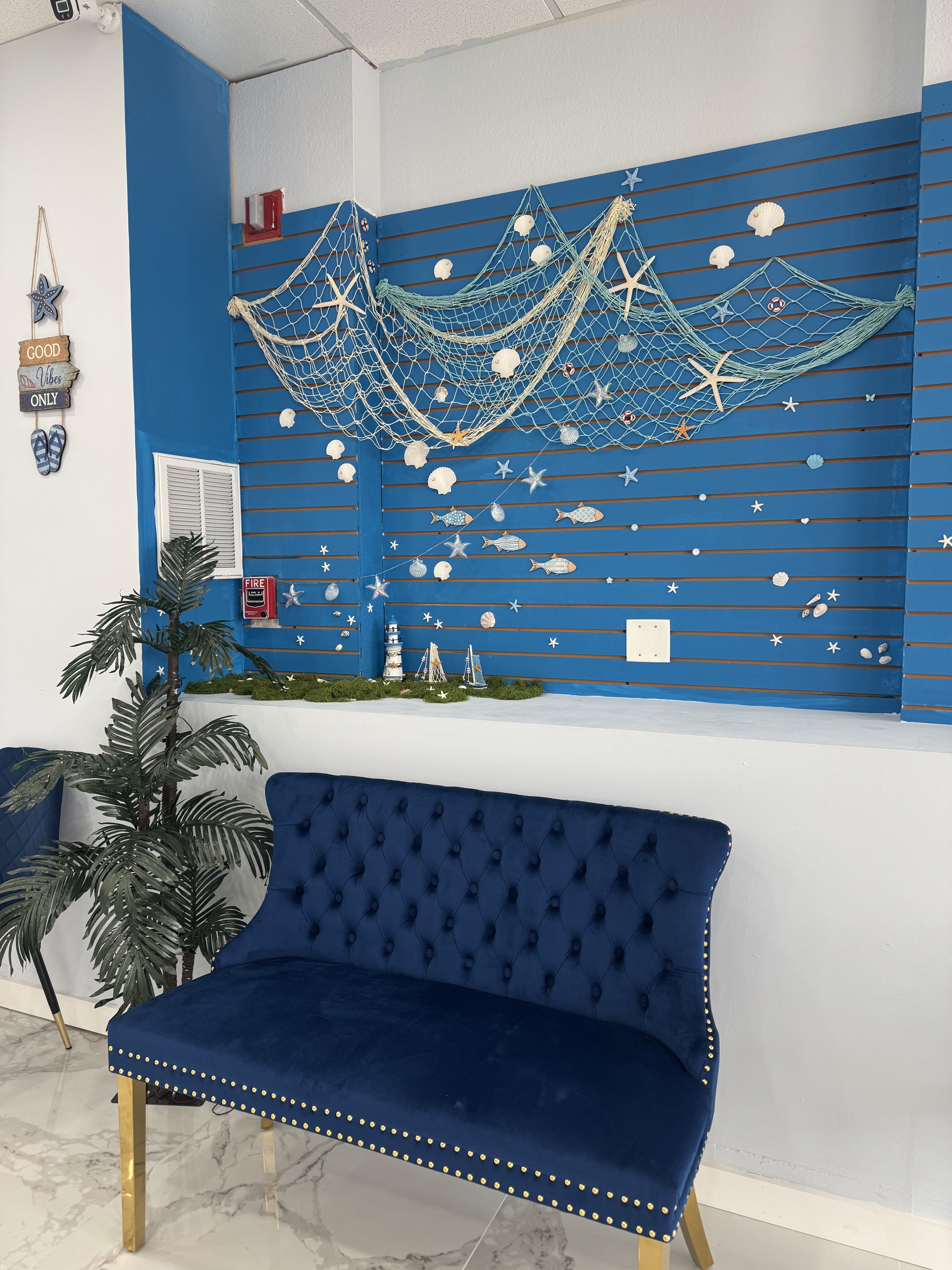 A blue velvet tufted bench with gold legs and studded detailing, placed against a white wall. To its left, there is a potted plant with broad leaves. Behind the bench, a blue slatted wall features a decorative fishing net with seashells, starfish, an