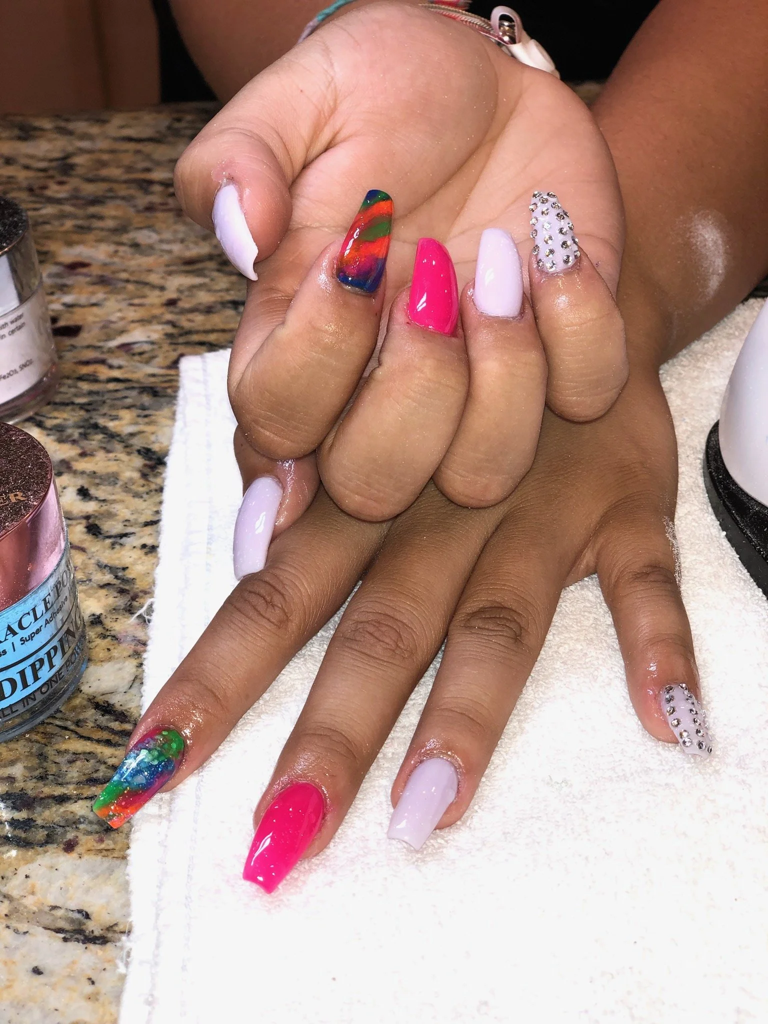 Close-up of a person's hands with colorful, decorated nails, resting on a white towel on a granite countertop, with nail polish bottles nearby.