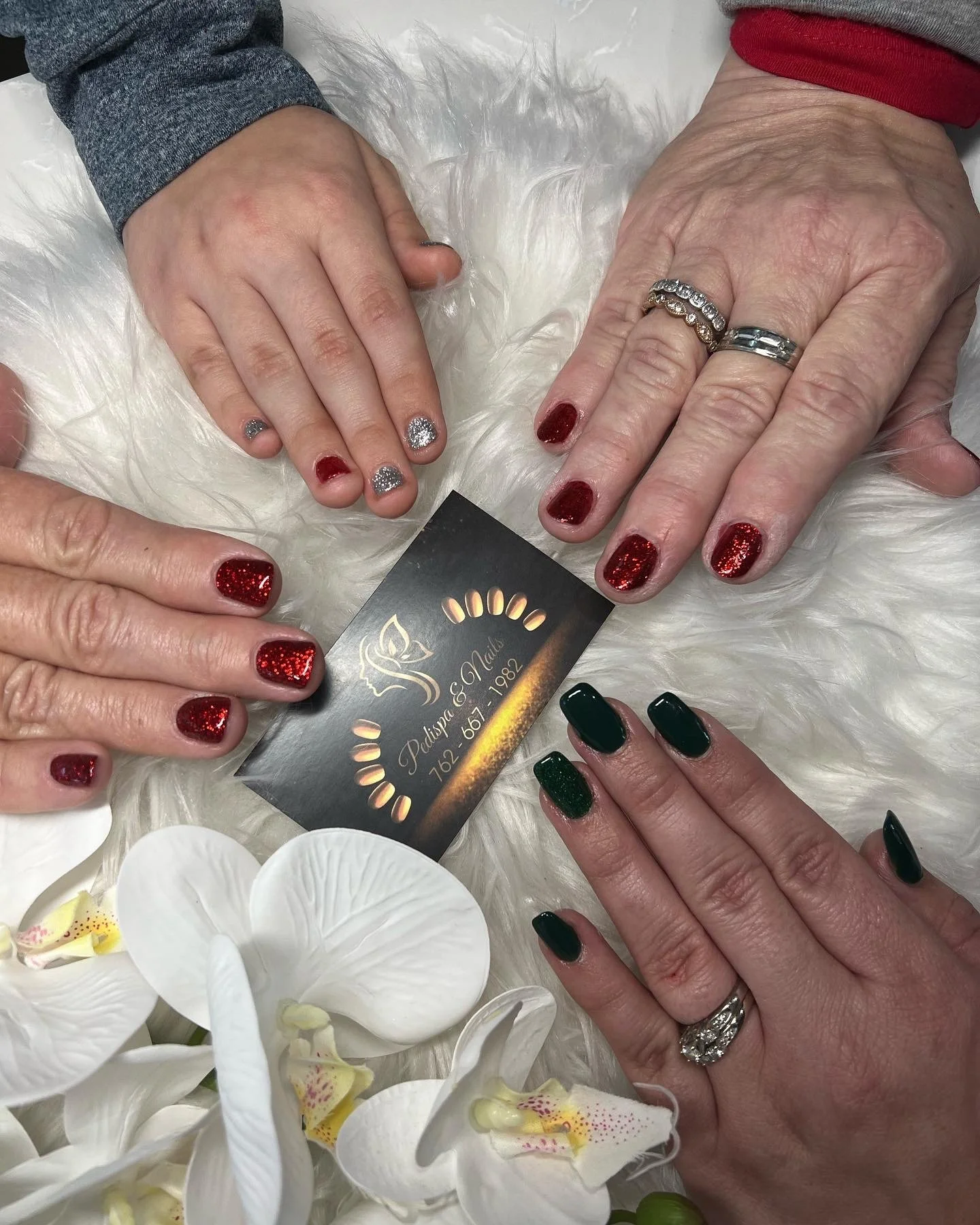 Close-up of four hands showing manicured nails, with a variety of glittery red, silver, and dark green nail polish, on a white furry surface surrounded by white orchids and a business card from a nail salon.