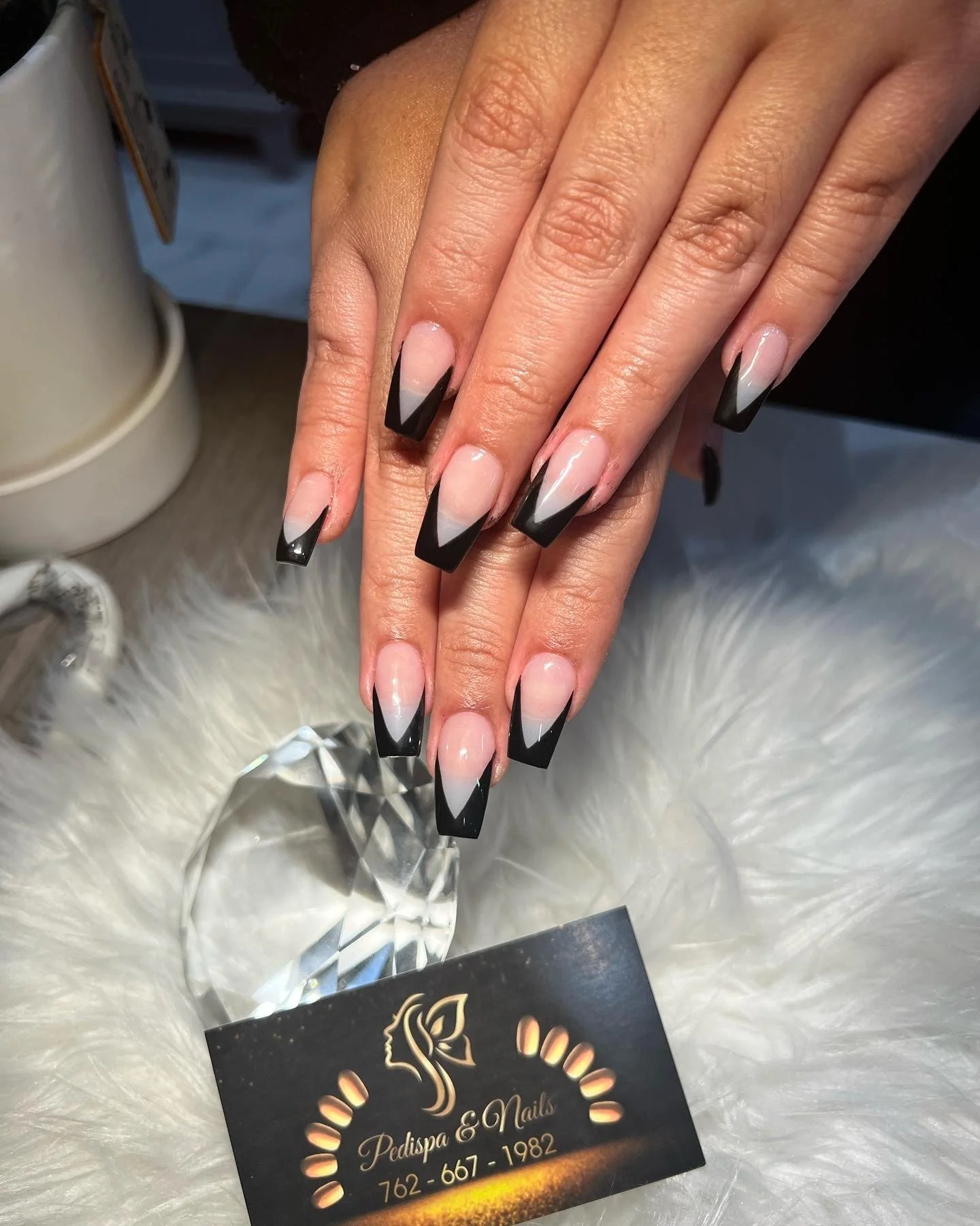 Close-up of hands with long square-shaped nails painted in a black and white geometric design, resting on a white furry surface with a large faceted crystal nearby and a business card for Pedispa & Nails underneath.