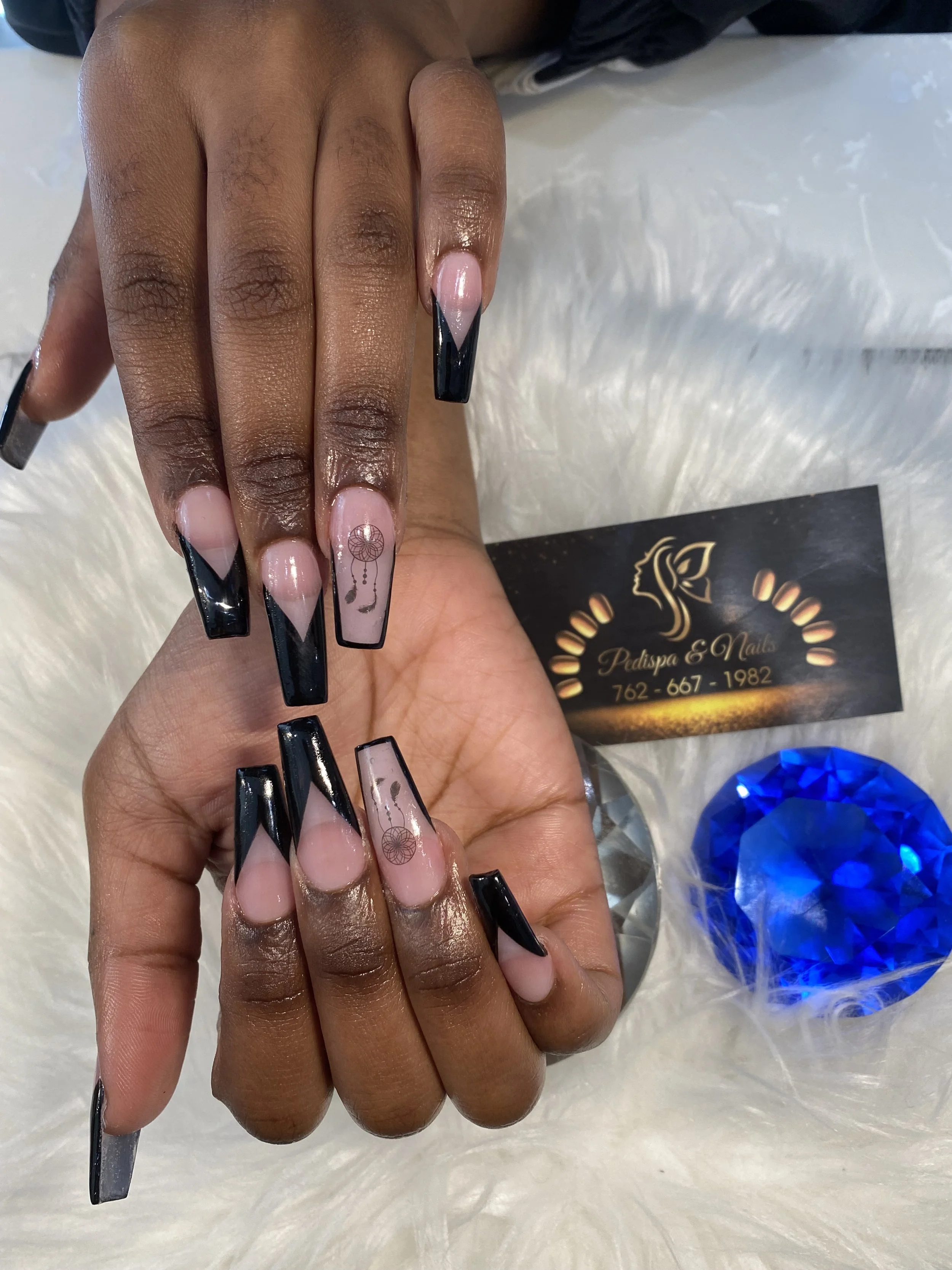 Close-up of a person's hand with long black coffin-shaped nails with a clear gloss and heart-shaped nail art. The hand is resting on a white surface with a business card and a large blue gem nearby.