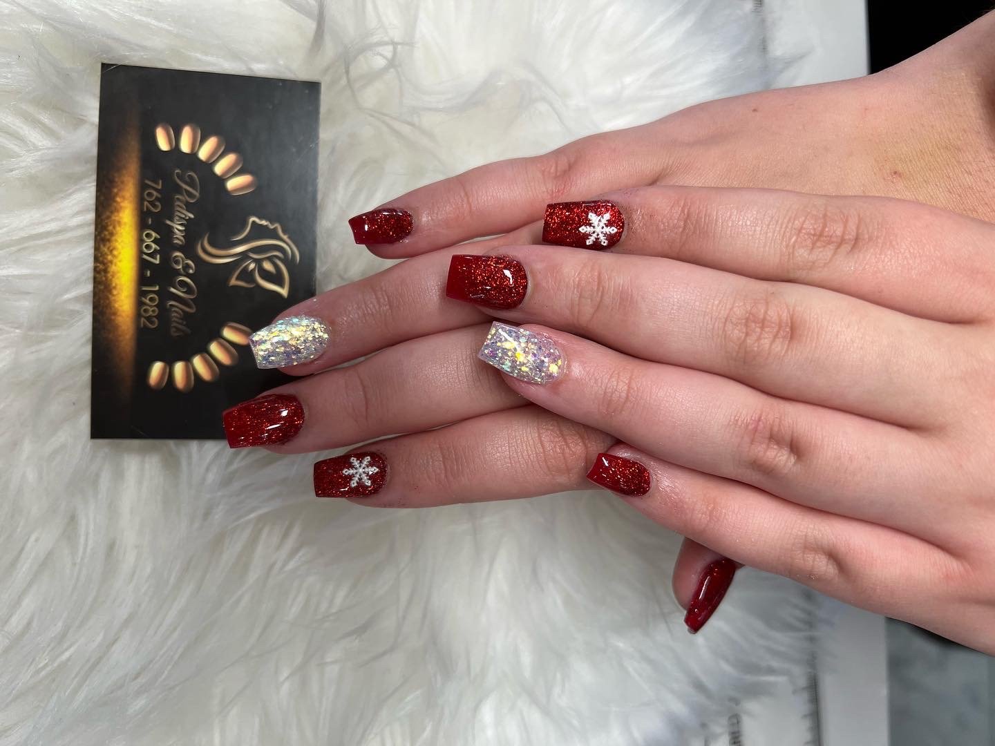 Hands with red and glittery nail polish, some nails have white snowflake designs, resting on a white furry surface next to a business card for a nail salon.