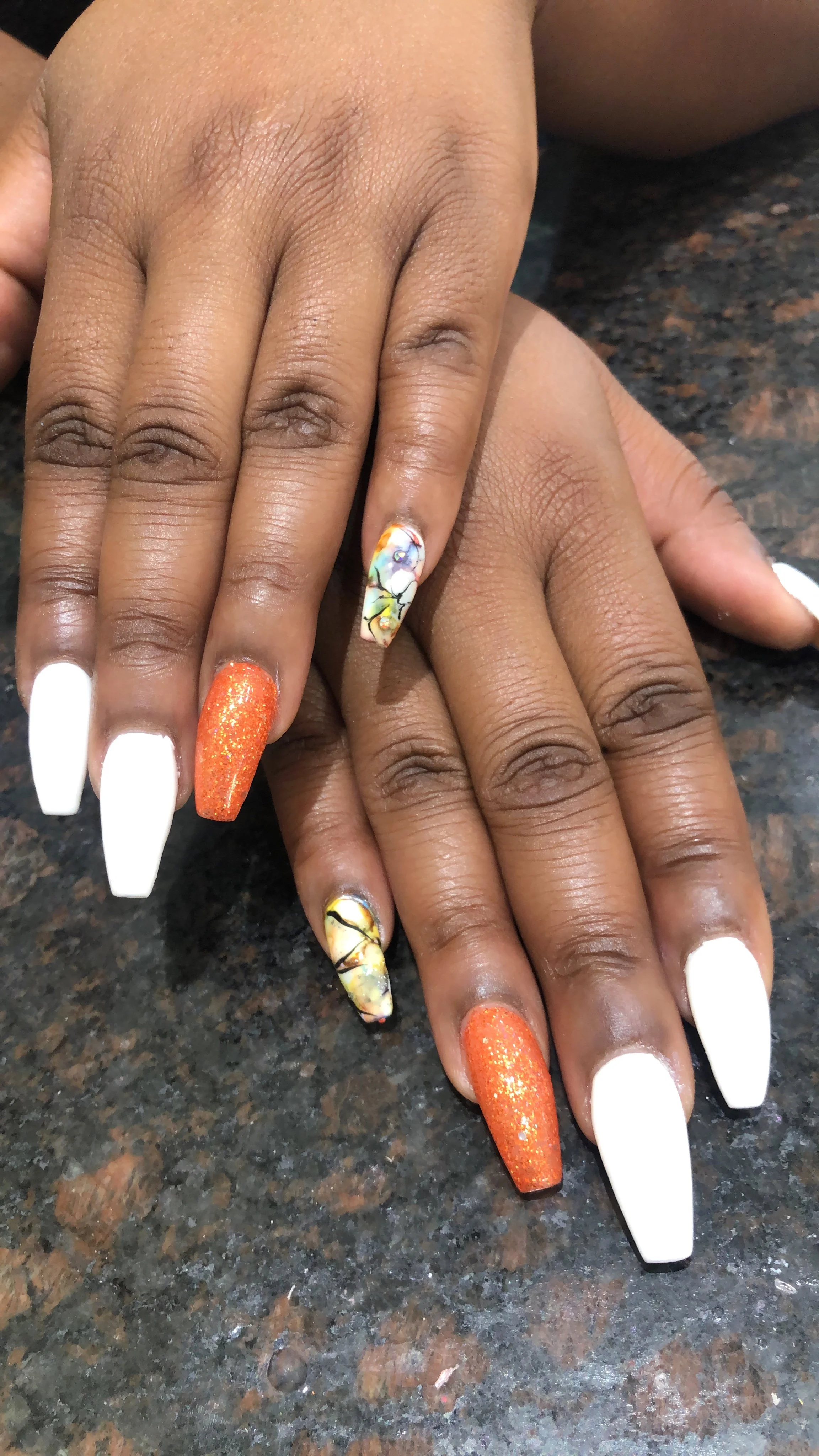 Close-up of hands with elaborate manicured nails, featuring a mix of white, orange glitter, and marble-like designs, resting on a dark, speckled surface.