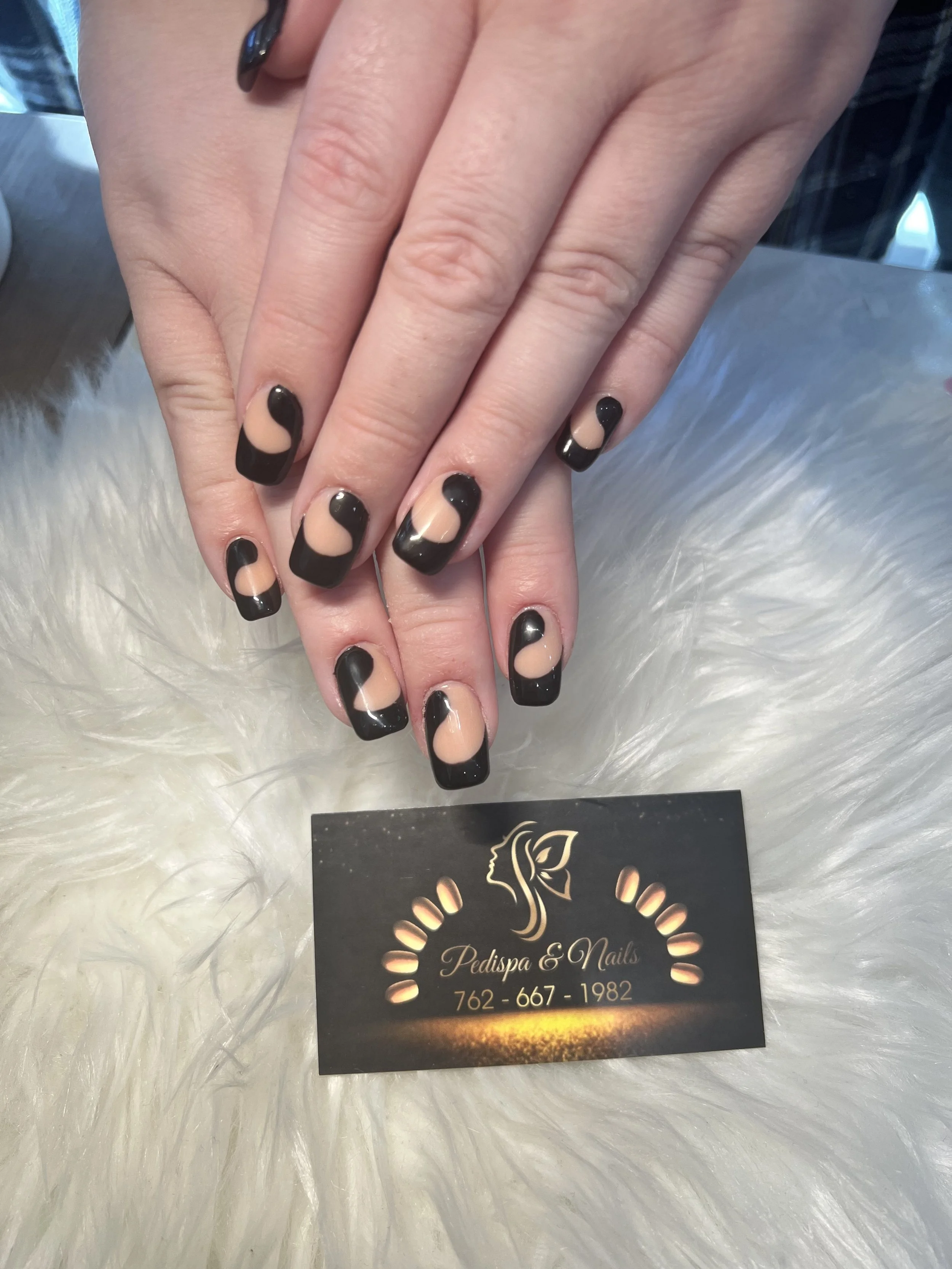 Close-up of a hand with black and nude French manicure nails, resting above a business card for Pedispa & Nails, on a white furry surface.