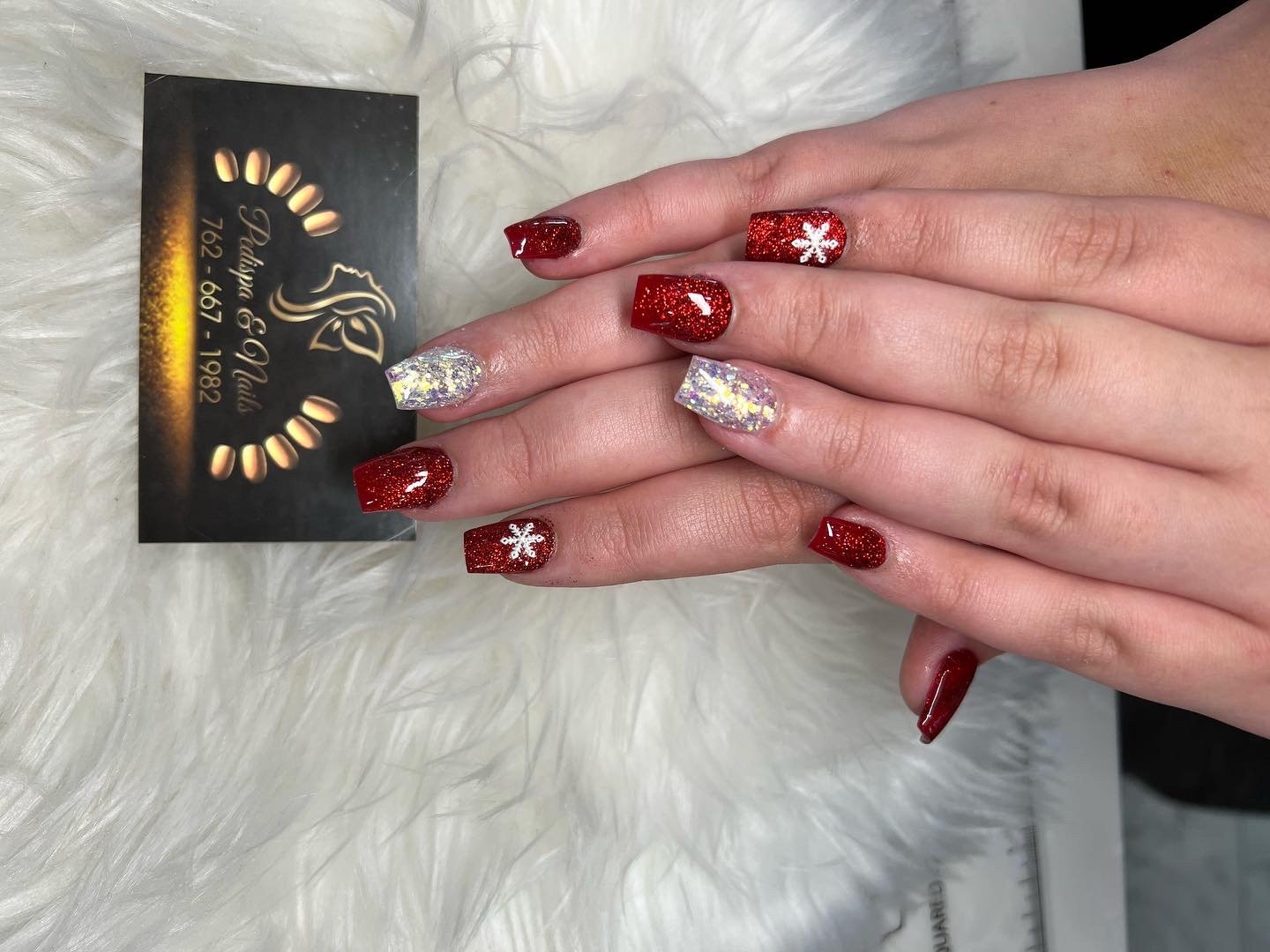 Hands with decorated nails resting on a white furry surface, with a black business card with gold lettering and a logo nearby. The nails are painted in red with glitter and white snowflake designs, and a couple are painted in holographic silver with 